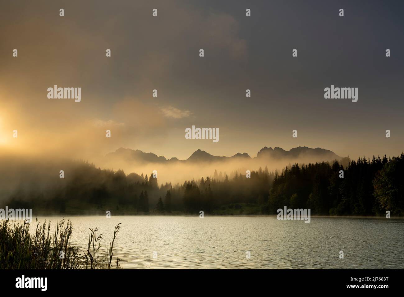 Nebel treibt trotz leichtem Wind kleine Wellen über den Geroldsee, auch Wagenbrüchsee genannt, in den bayerischen Alpen vor ihm. Die Sonne zaubert sanfte Farbtöne und im Hintergrund erstreckt sich das Karwendel. Stockfoto