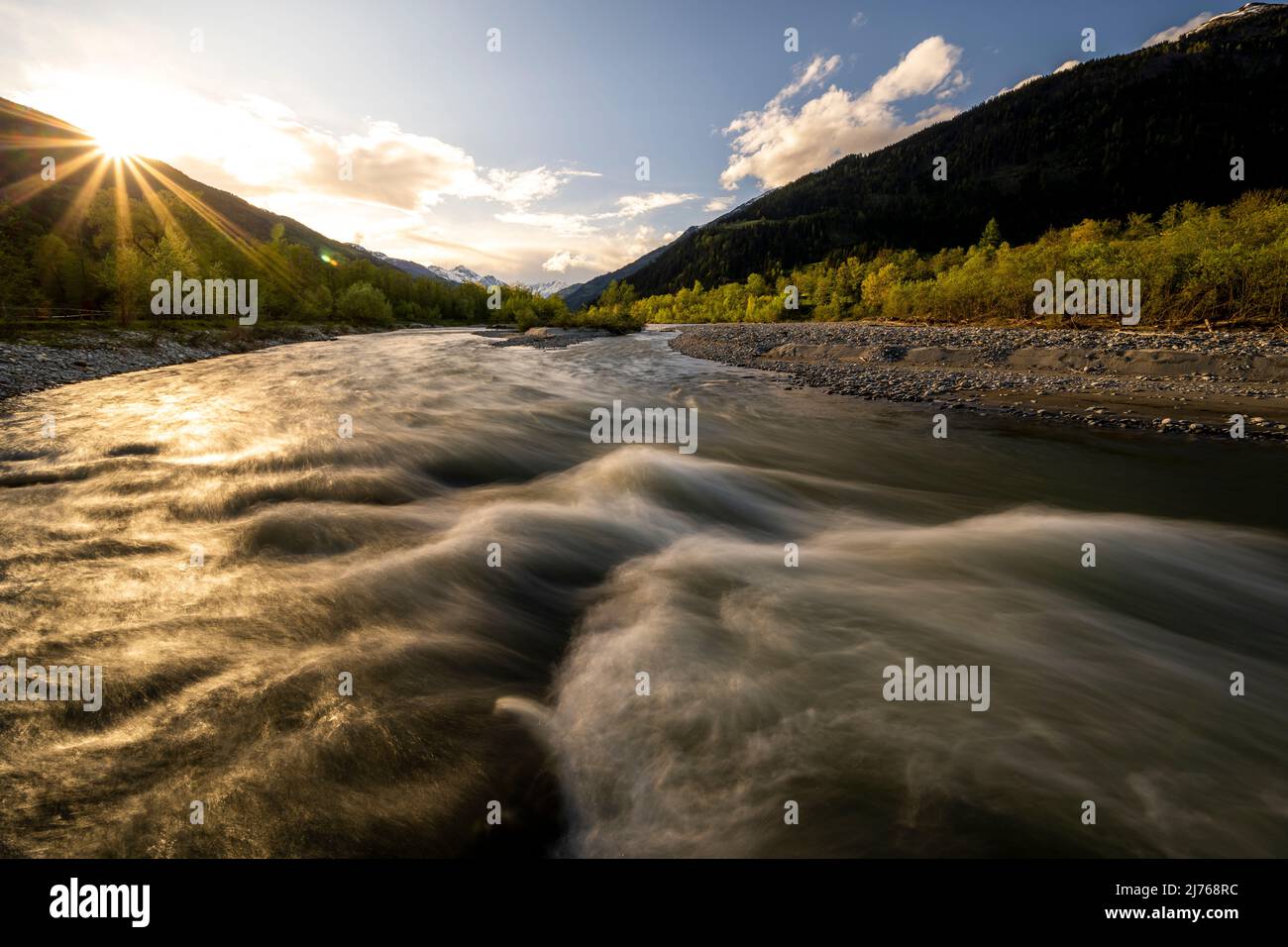 Sonnenuntergang mit Sonnenstern an der Isel, einem wilden Fluss in Osttirol bei Lienz, mit Stromschnellen am Fließwasser. Stockfoto