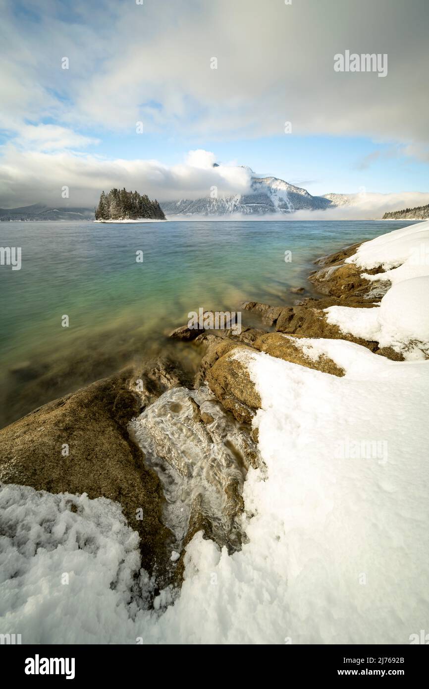 Winter mit Schnee, Wolken und Nebel am Walchensee, im Hintergrund die Insel Sassau und Herzogstand in den bayerischen Alpen. Das türkisgrüne Wasser bildet an der felsigen Küste etwas Eis. Stockfoto