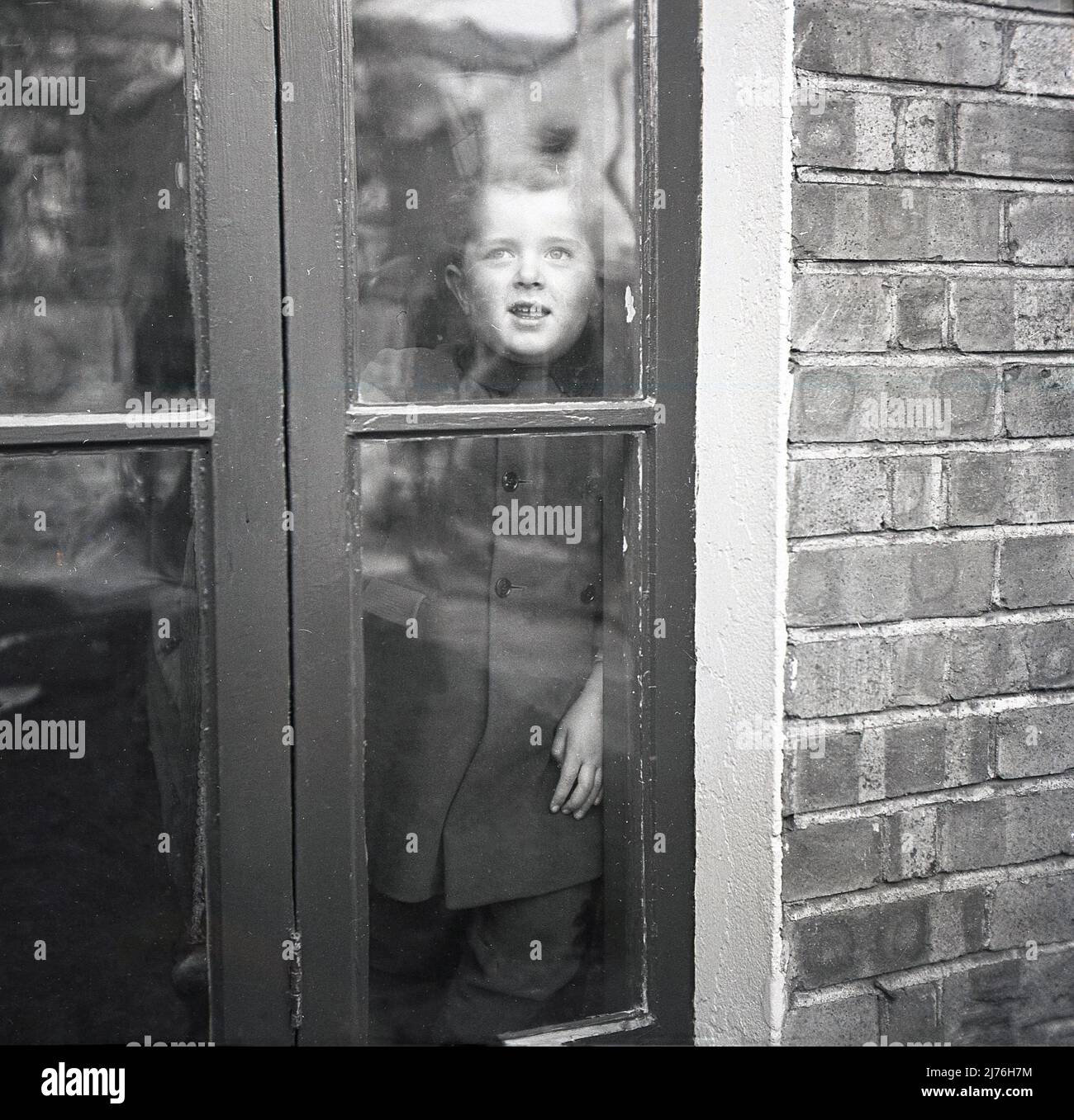 1950s, historisch, ein kleiner Junge im Inneren, der an hölzernen französischen Türen stand und nach draußen schaute, England, Großbritannien. Stockfoto