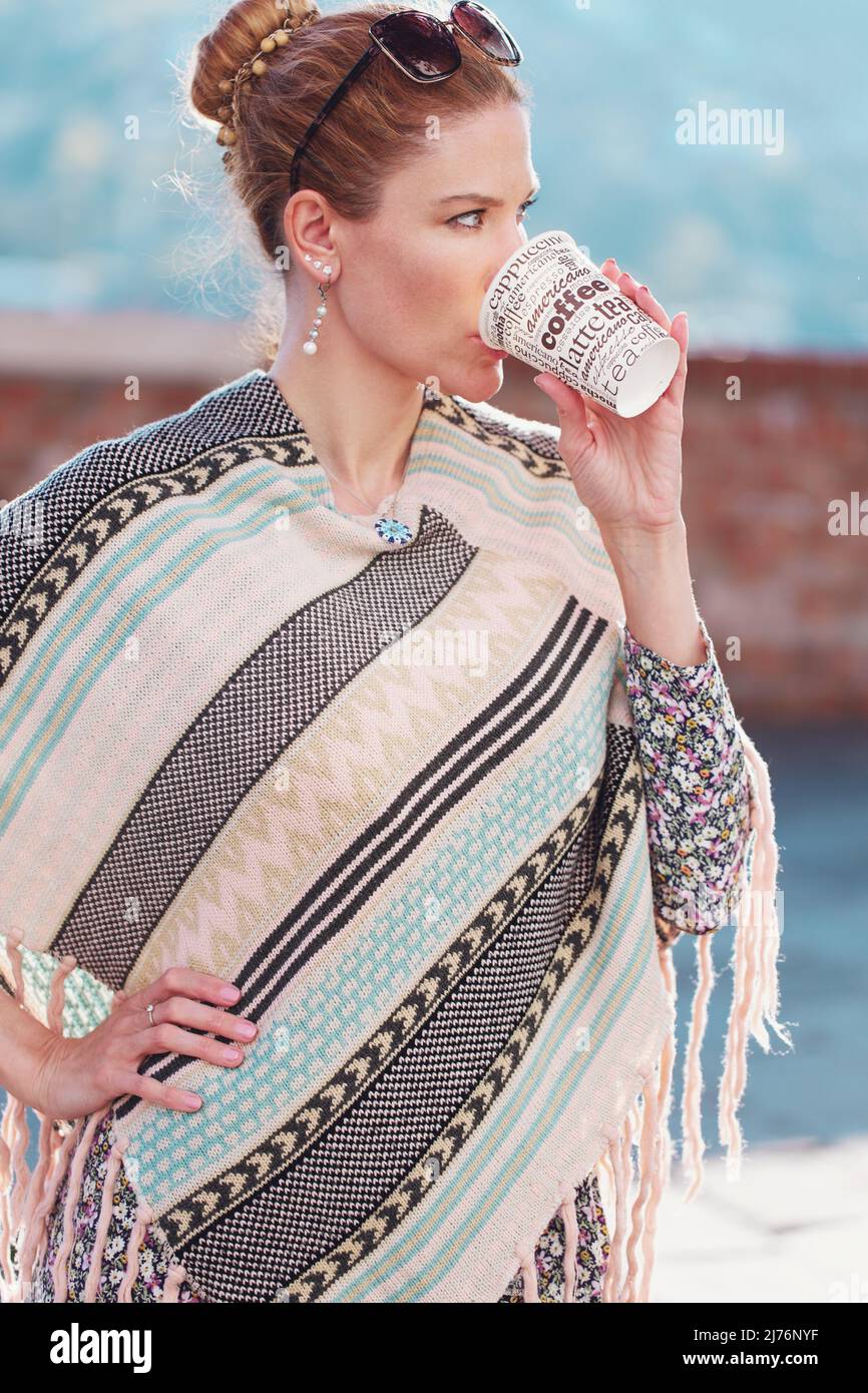 Junge fröhliche Rothaarige Frau in Poncho trinkt Kaffee im Freien, vertikal Stockfoto