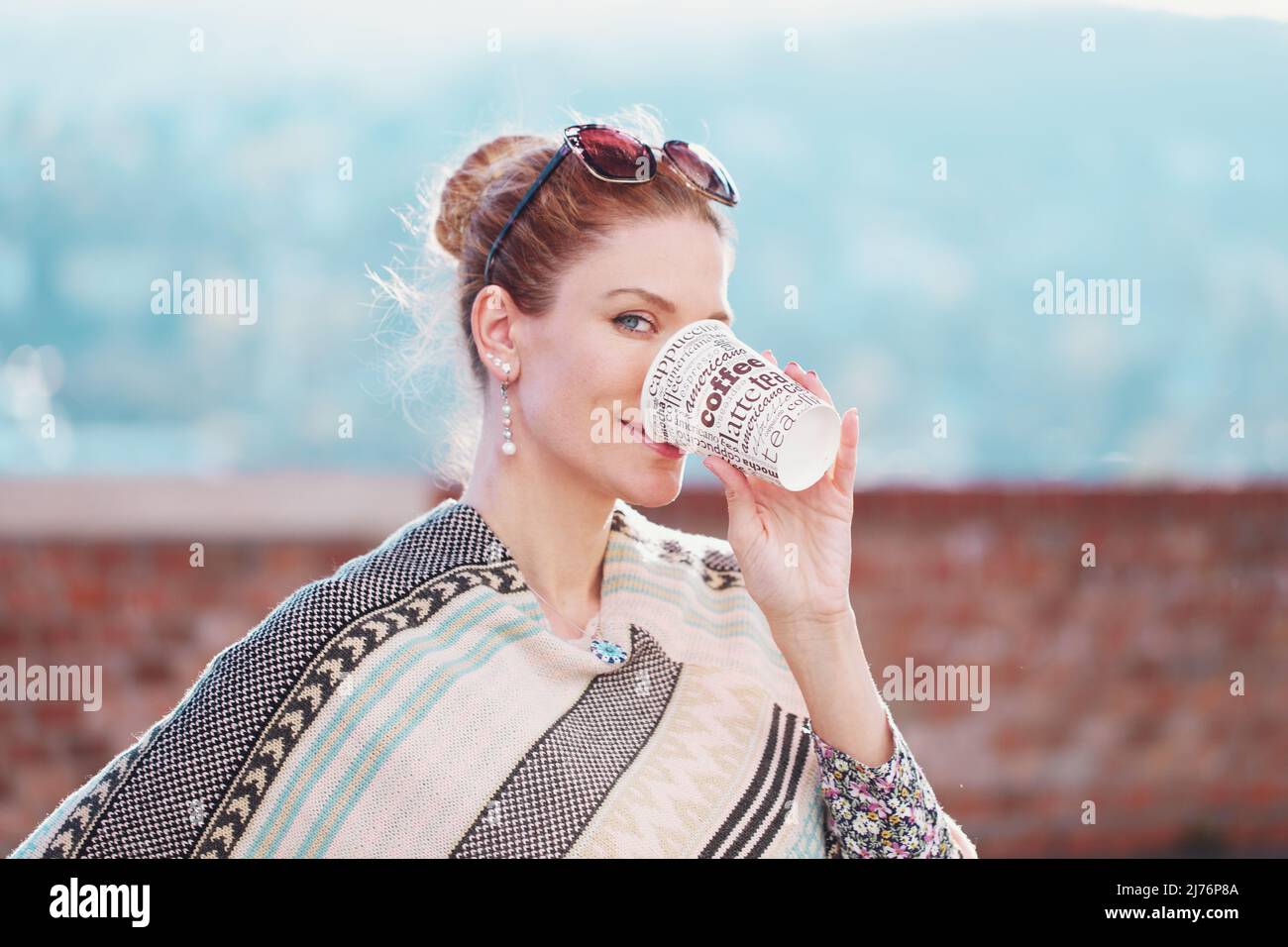Junge fröhliche Rothaarige Frau im Poncho, die im Freien Kaffee trinkt und in die Kamera schaut Stockfoto