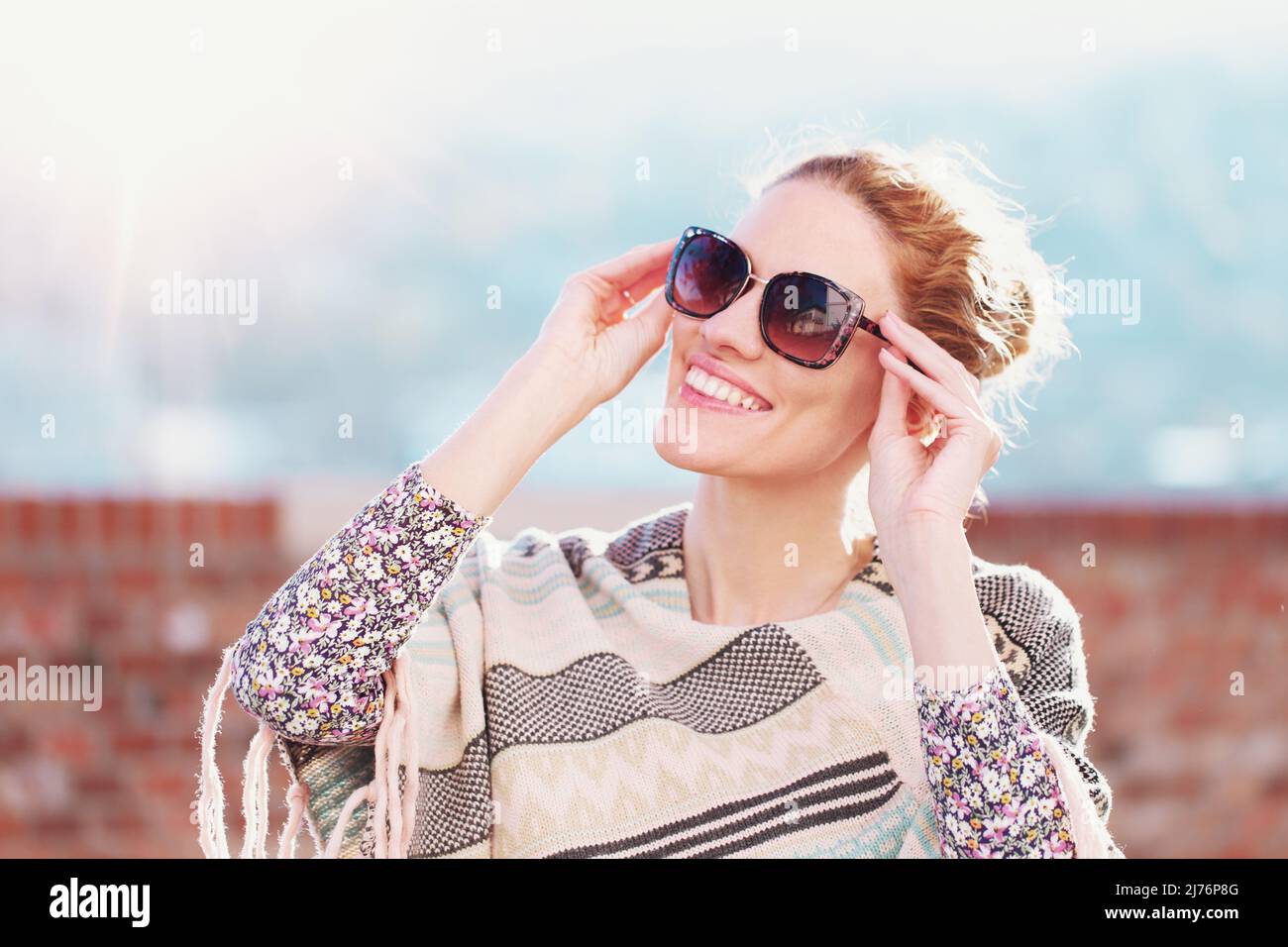 Fröhliche junge Rotschopf-Frau, die draußen Sonnenschein genießt und eine Sonnenbrille hält Stockfoto