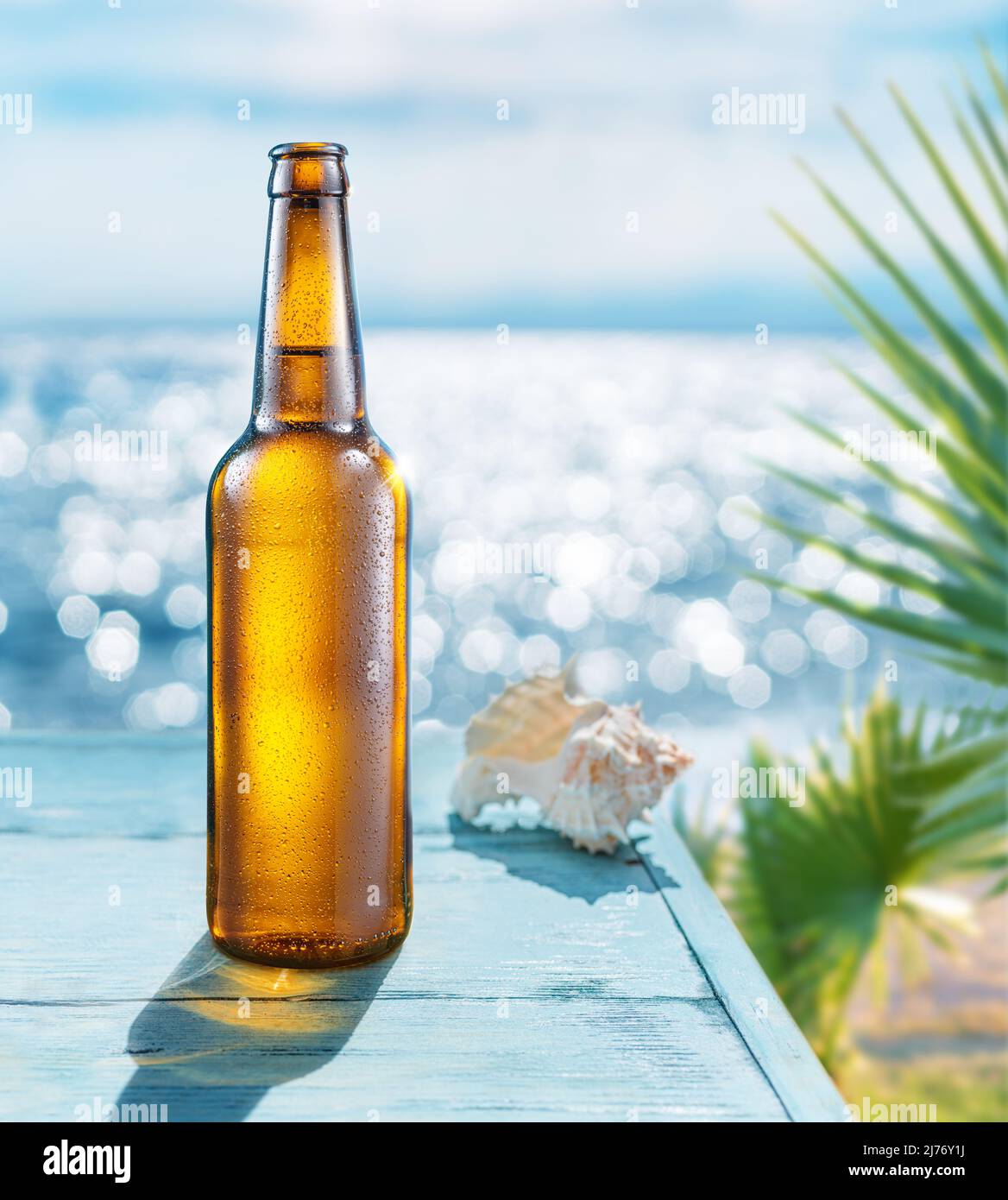 Geöffnete Flasche Bier mit Kondenswasser auf dem Holztisch. Verschwommenes glitzerndes Meer im Hintergrund. Sommerliche Stimmung. Stockfoto