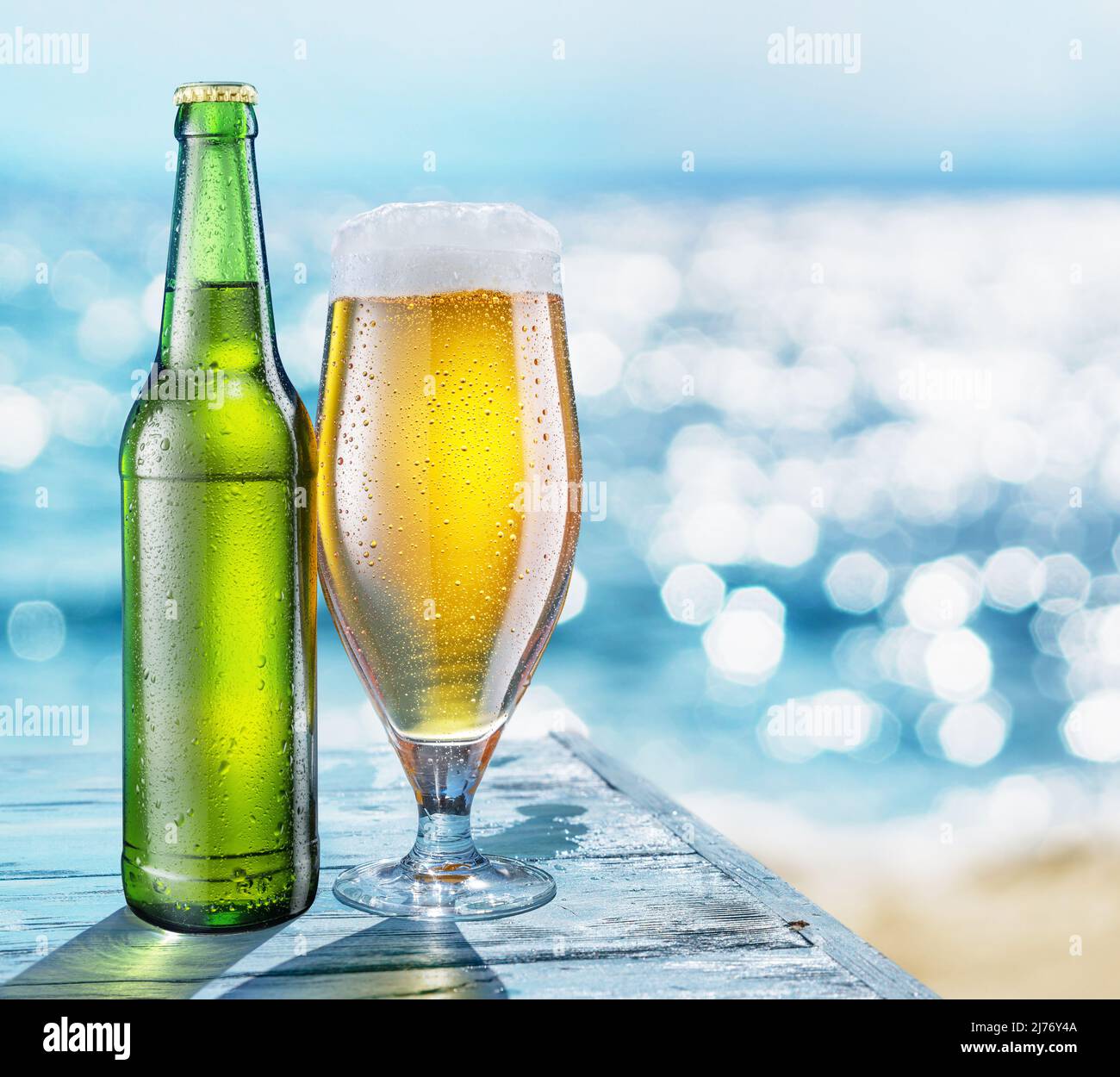 Flasche Bier und Bierglas mit Kondenswasser auf dem Holztisch. Verschwommenes glitzerndes Meer im Hintergrund. Sommerliche Stimmung. Stockfoto