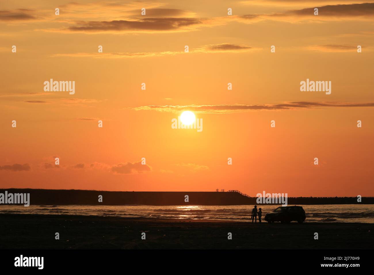 Ein japanisches Paar, das an einem Sandstrand steht, an dem die Sonne im Meer untergeht Stockfoto