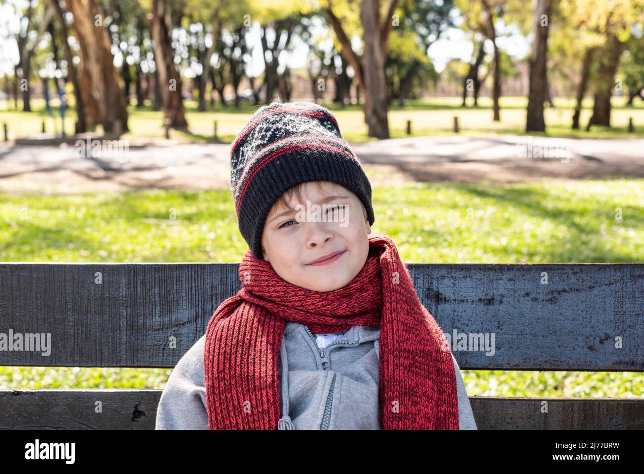 Das Kind sitzt auf einer Bank im Park und trägt einen Wollhut und einen Schal Stockfoto