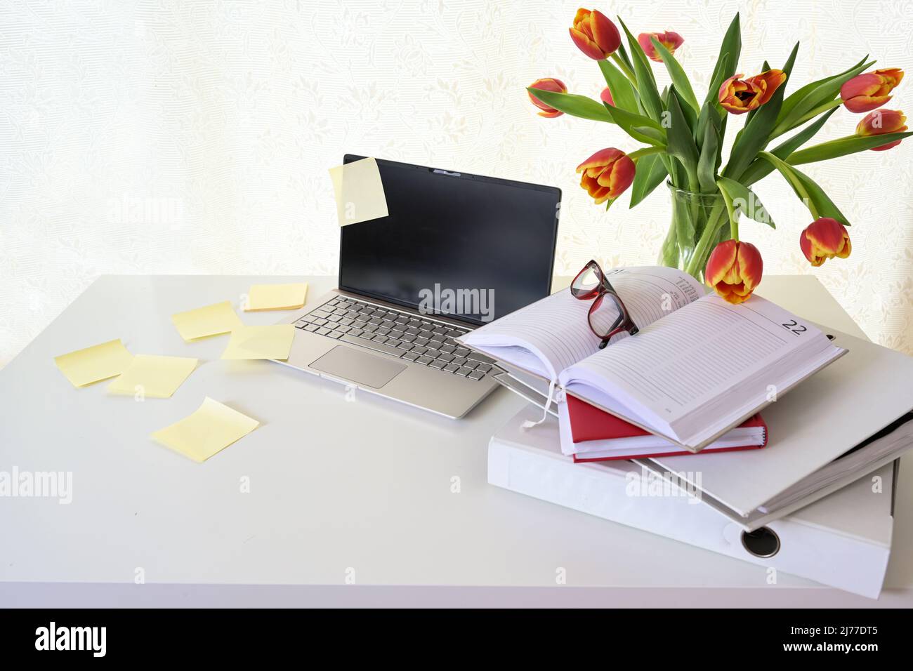 Laptop mit vielen Haftnotizen, Ringordner, Organizer und einem Strauß roter Tulpen auf einem weißen Schreibtisch, Konzept für Stress im Home Office, Business oder Stockfoto