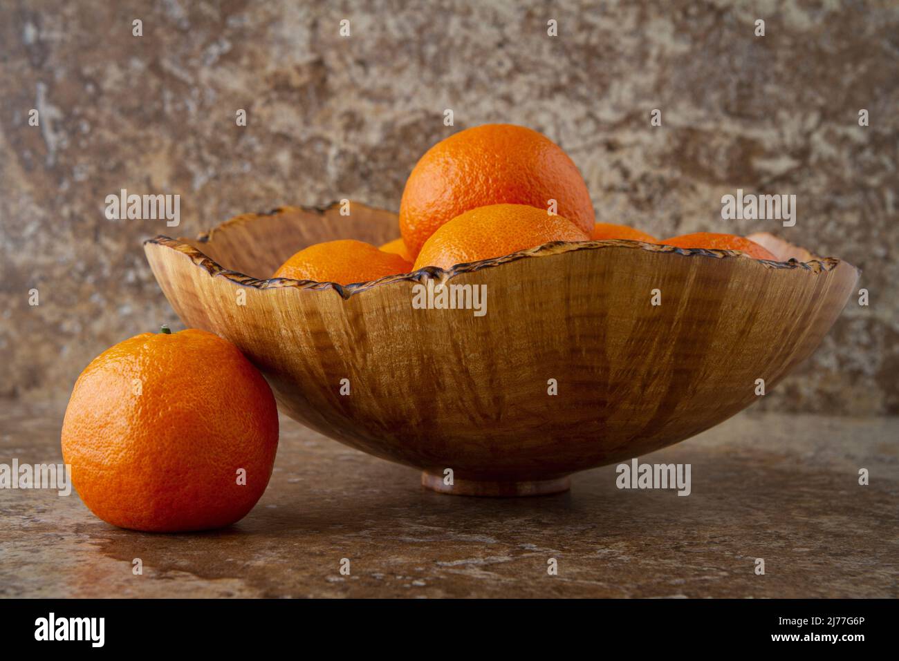 Ein Stillleben einer dekorativen Holzschale gefüllt mit Mandarinen auf warm getönten braunen Fliesen mit einer einzigen Frucht neben der Schale. Stockfoto