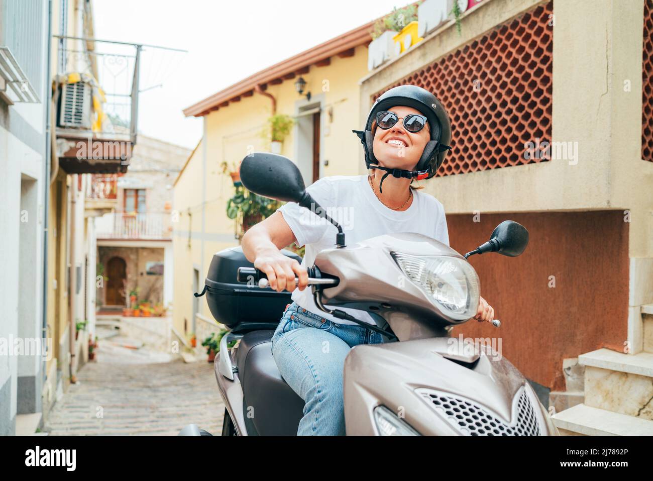 Fröhlich lächelnde Frau mit Helm und Sonnenbrille fährt schnell mit dem Moto Scooter auf den engen Straßen der sizilianischen Altstadt. Glücklicher italienischer Urlaub und tra Stockfoto