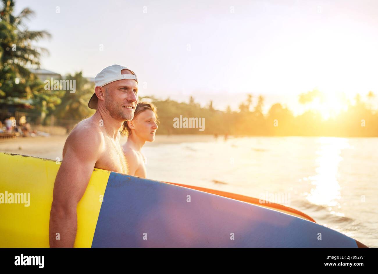 Lächelnder Mann mit jungendlichen Jungen Sohn mit Surfbrettern geht zum Surfen ins Meer. Sie haben einen Winterurlaub und genießen ein wunderschönes Sonnenuntergangslicht. Familie Stockfoto