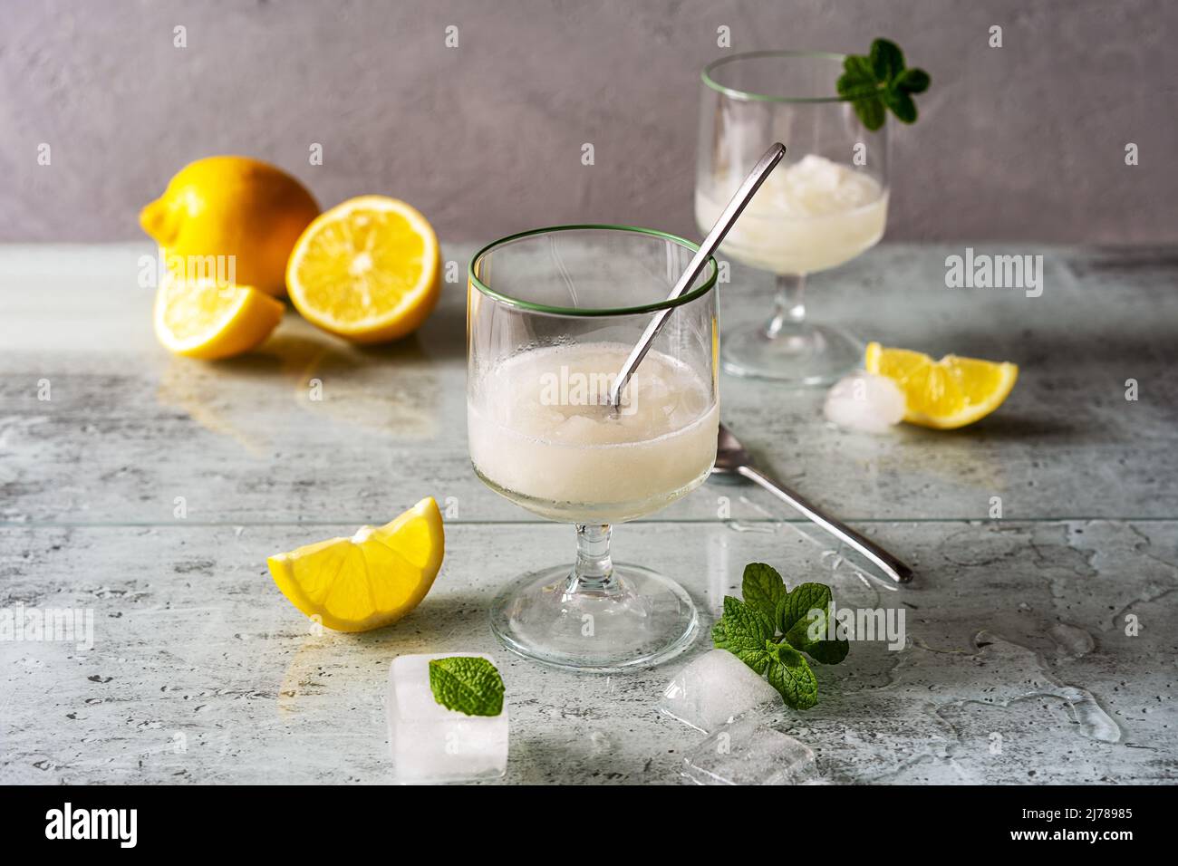 Zitronensorbet in zwei Gläsern mit Zitronen, Eiswürfeln, Teelöffeln, Minzblättern auf grauem Hintergrund Stockfoto