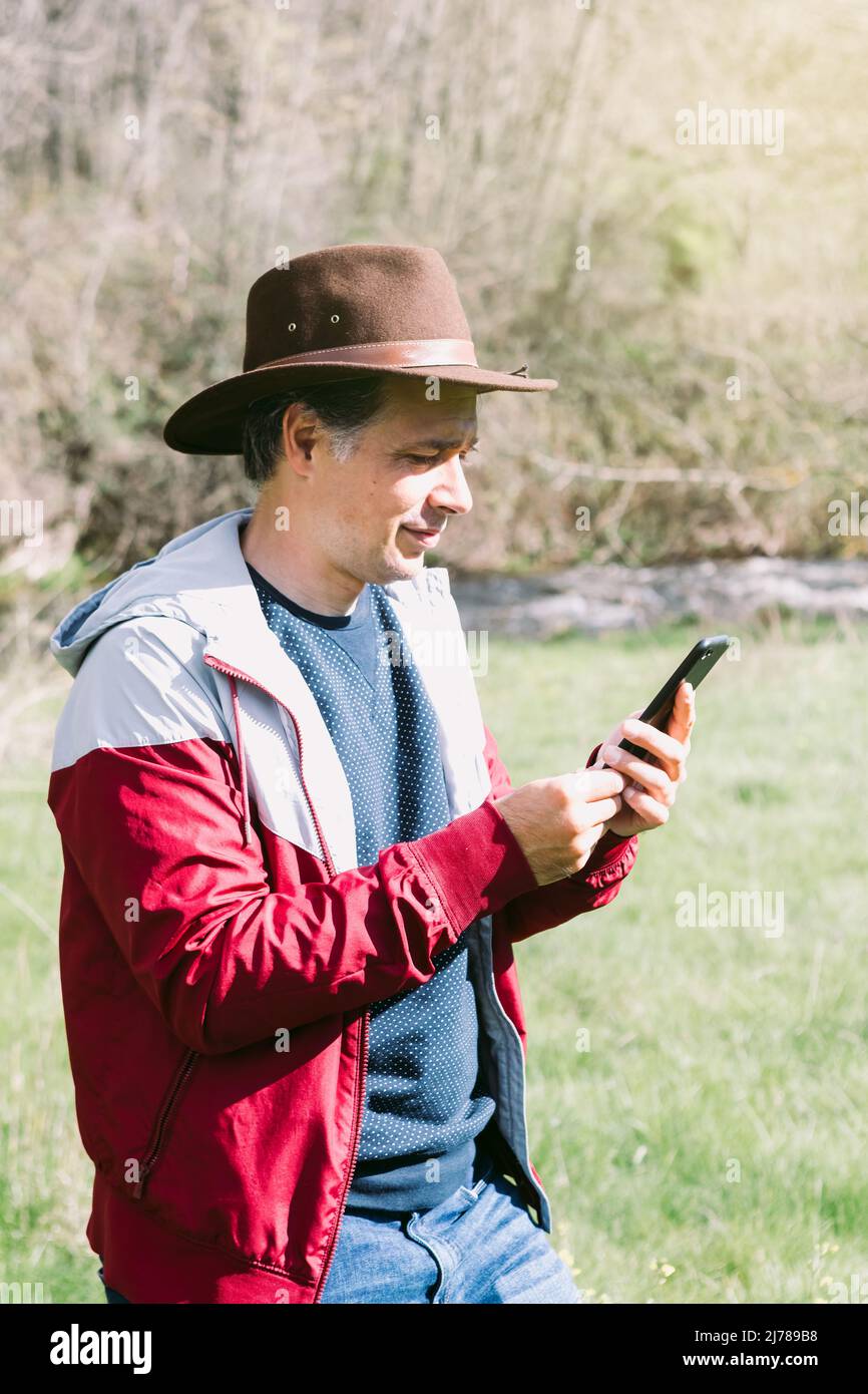 Unternehmerischer, selbstständiger Mann mit Hut, der auf sein Mobiltelefon blickt, während er sich auf dem Feld auf einer Wiese entspannt. Konzept der Arbeit, genießen, entspannen, Stockfoto