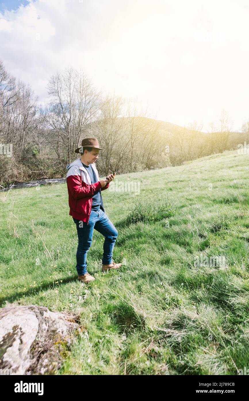 Unternehmerischer, selbstständiger Mann mit Hut, der auf sein Mobiltelefon blickt, während er sich auf dem Feld auf einer Wiese entspannt. Konzept der Arbeit, genießen, entspannen, Stockfoto