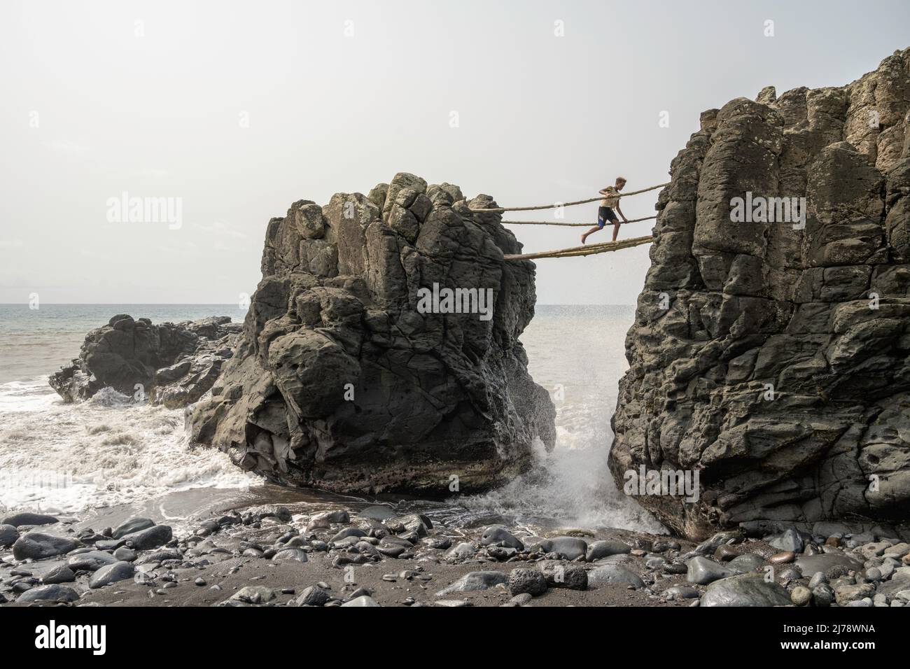 Junge überquert eine prekäre Brücke aus Baumstämmen, während die Wellen auf die Felsen schlagen. Stockfoto