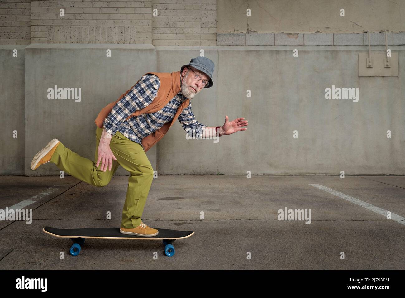 Bärtiger älterer Mann mit runder Brille und einem Eimer Hut reitet auf einem langen Skateboard in einer grunge städtischen Umgebung Stockfoto