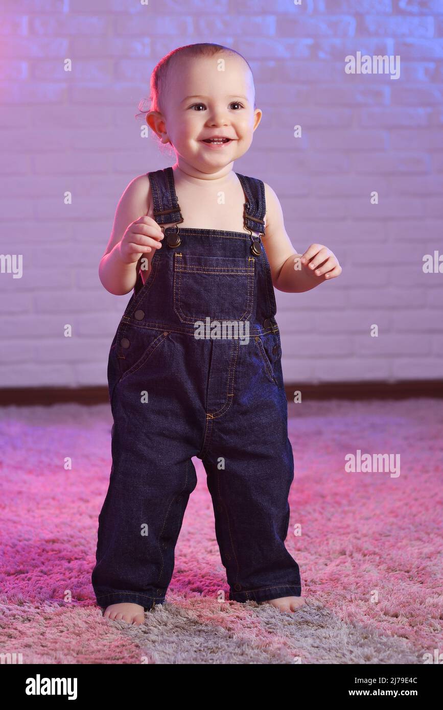 Baby Boy in Denim Overalls lächelnd, während er barfuß auf dem Teppich vor dem Hintergrund einer weißen Ziegelwand steht Stockfoto