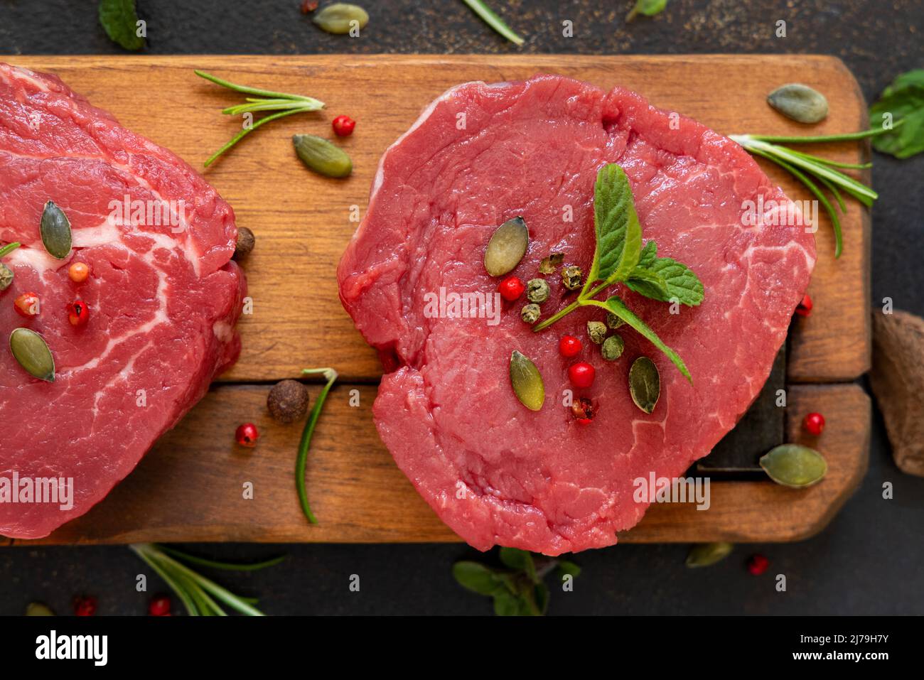 Rohe Rinderfiletsteaks auf Holzbrett auf dunklem Tisch aus nächster Nähe. Portionierte Filet-Mignons mit Gewürzen und Kräutern. Konzept der Sommerkost Stockfoto