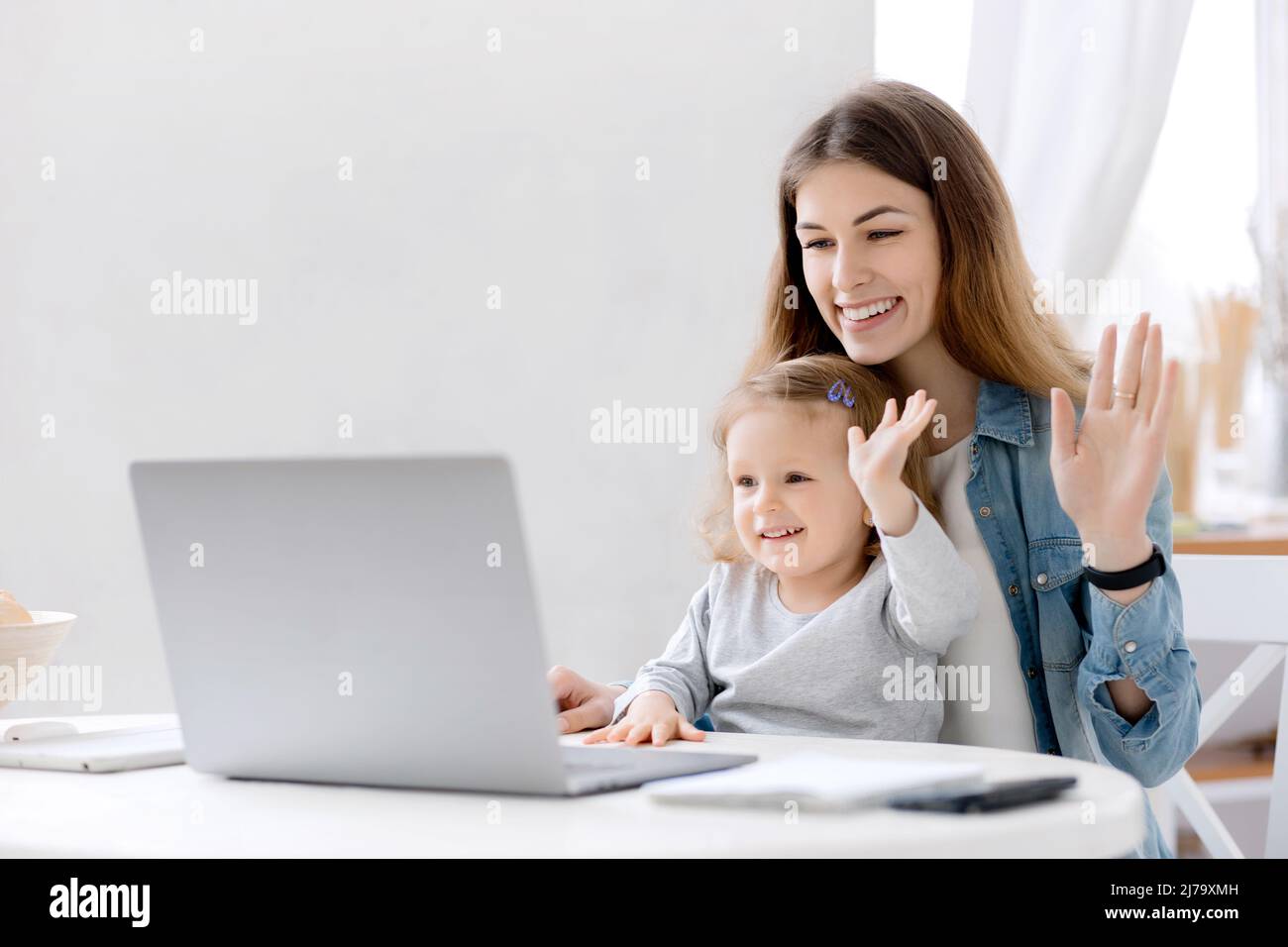 Schöne Mutter und kleines Baby arbeitet von zu Hause aus, sie sitzt in der Küche und mit Laptop für Remote-Arbeit, sitzt ihr nettes kleines Mädchen in der Nähe auf einem Stockfoto