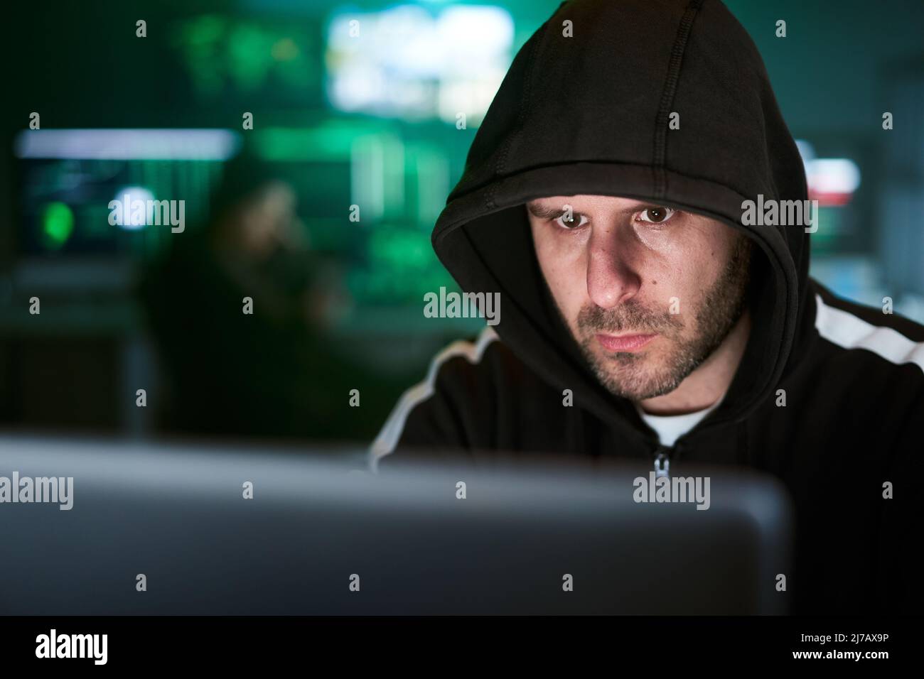 Porträt eines geheimnisvollen, mit Kapuze versehenen Hackers, der am Schreibtisch sitzt und in dunkler Atmosphäre in die Datenserver der Regierung einbricht Stockfoto