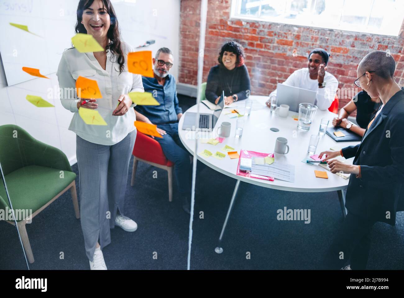 Lächelnde Geschäftsfrau, die sich mit ihren Kollegen im Hintergrund an einer Glaswand Notizen macht. Kreative junge Geschäftsfrau, die ihre Ideen mit ihrem Team teilt Stockfoto