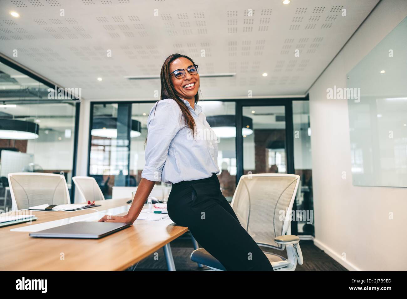 Geschäftsfrau lächelt fröhlich in einem Sitzungssaal. Geschäftsfrau im mittleren Erwachsenenalter, die sich an einem modernen Arbeitsplatz an einem Tisch lehnt. Fröhliche Unternehmerin W Stockfoto