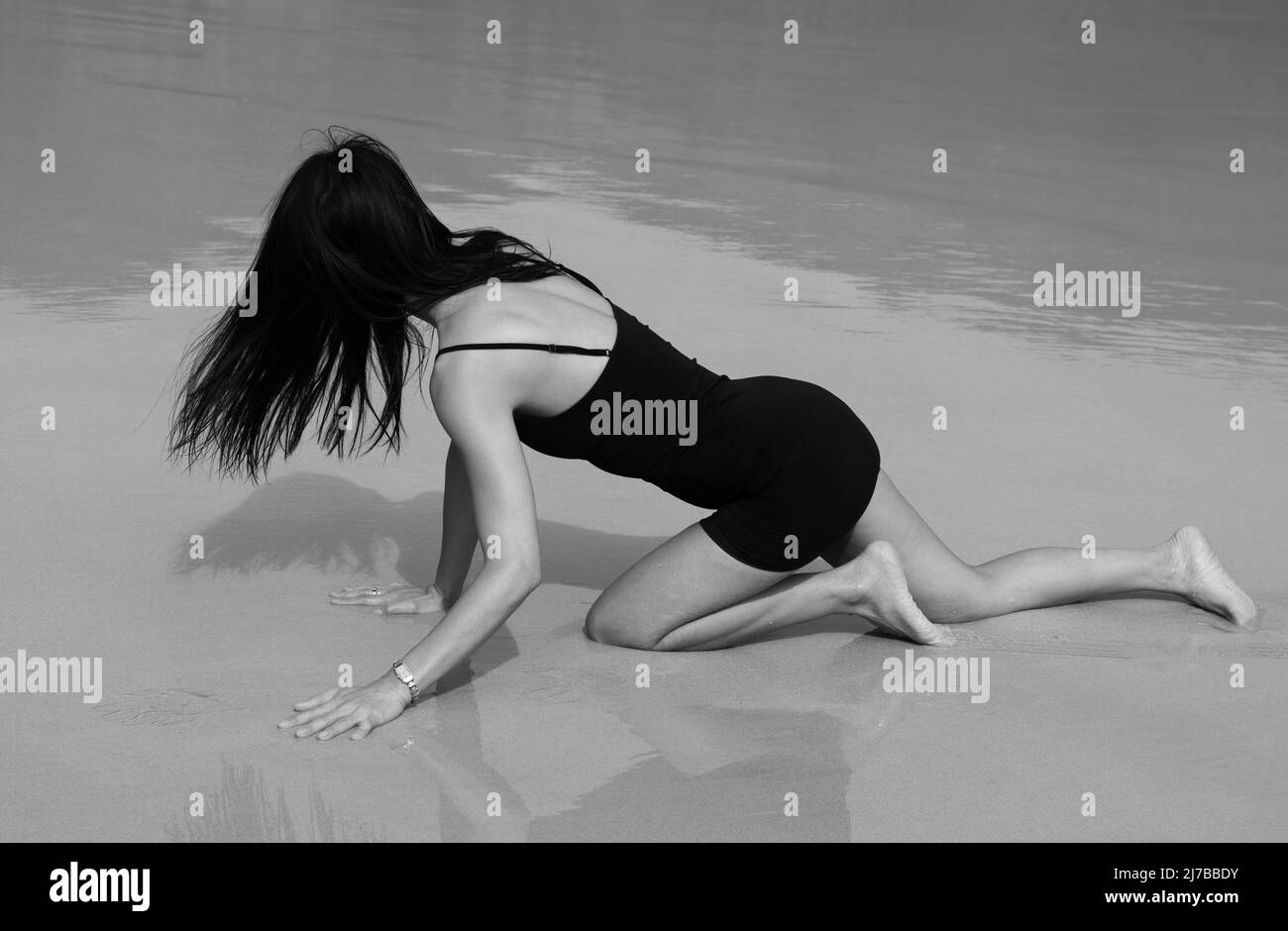 Eine Frau in einem kurzen schwarzen Kleid mit langen schwarzen Haaren kriecht auf nassem Sand am Strand. Ihr Schatten und ihre Reflexion sind zu sehen. Monochrom. Stockfoto