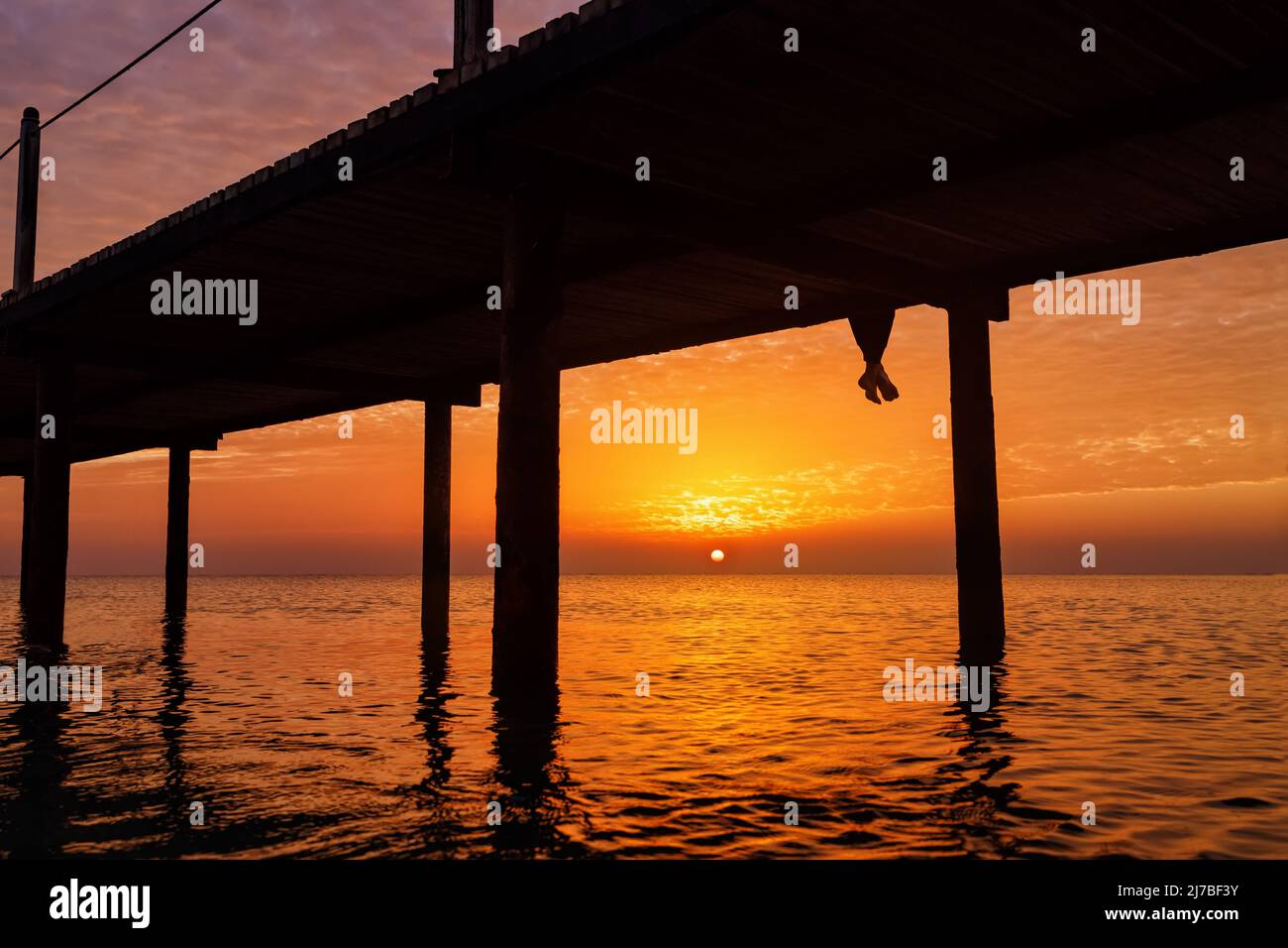 Beine einer Frau, die bei Sonnenaufgang auf einer Holzbrücke mit wunderschöner Meereslandschaft sitzt Stockfoto