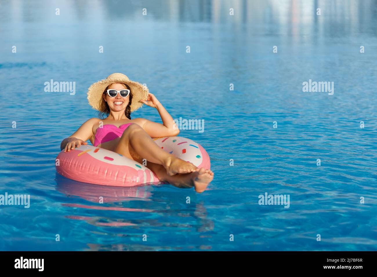 Lächelnde junge Frau in Strohhut und Sonnenbrille, die sich auf einem aufblasbaren Ring im Schwimmbad entspannt Stockfoto