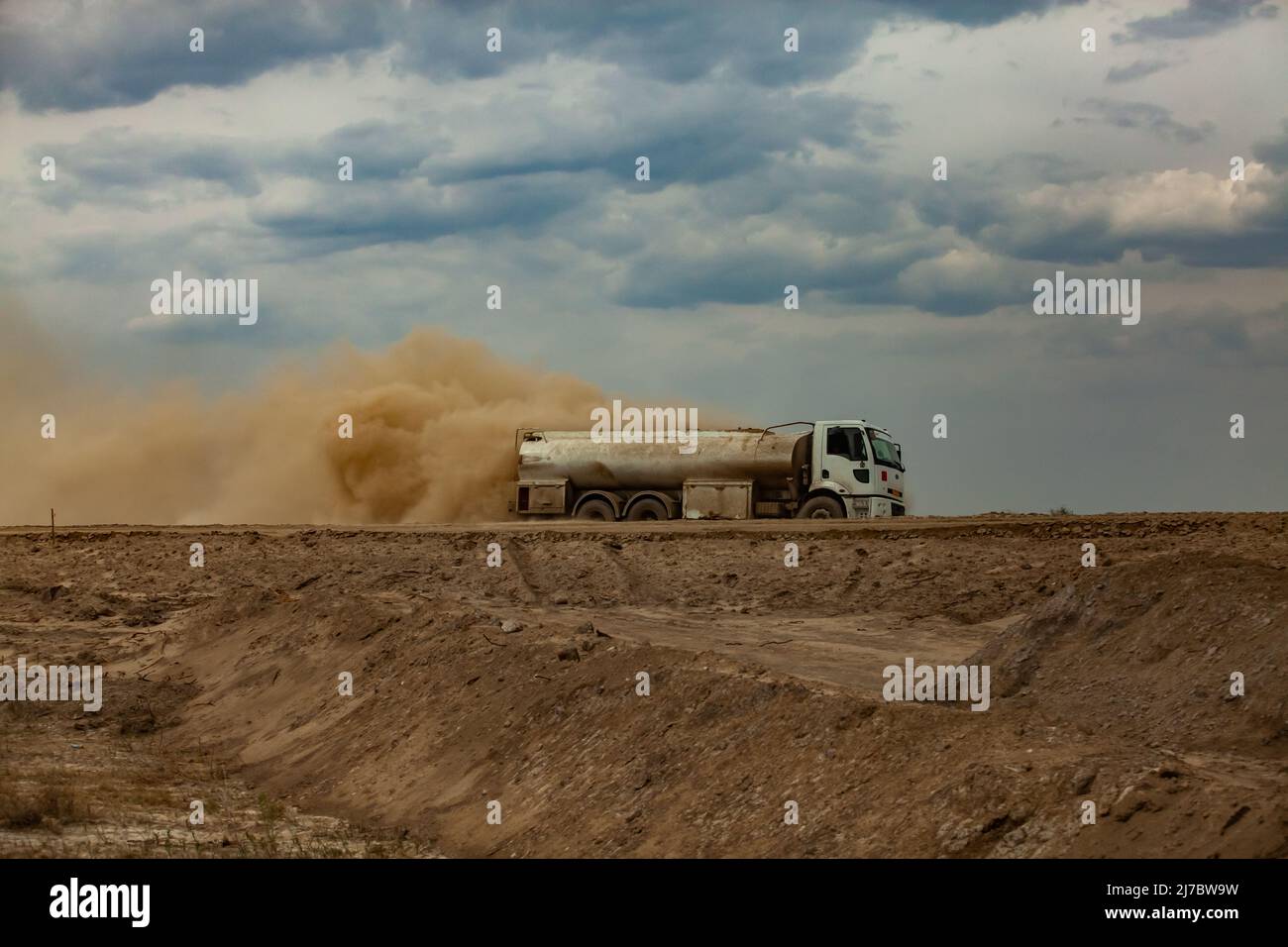 Tankwagen und gelbe Staubwolke auf Sandstraße gegen grau bewölkten Himmel. Straßenbaustelle. Stockfoto