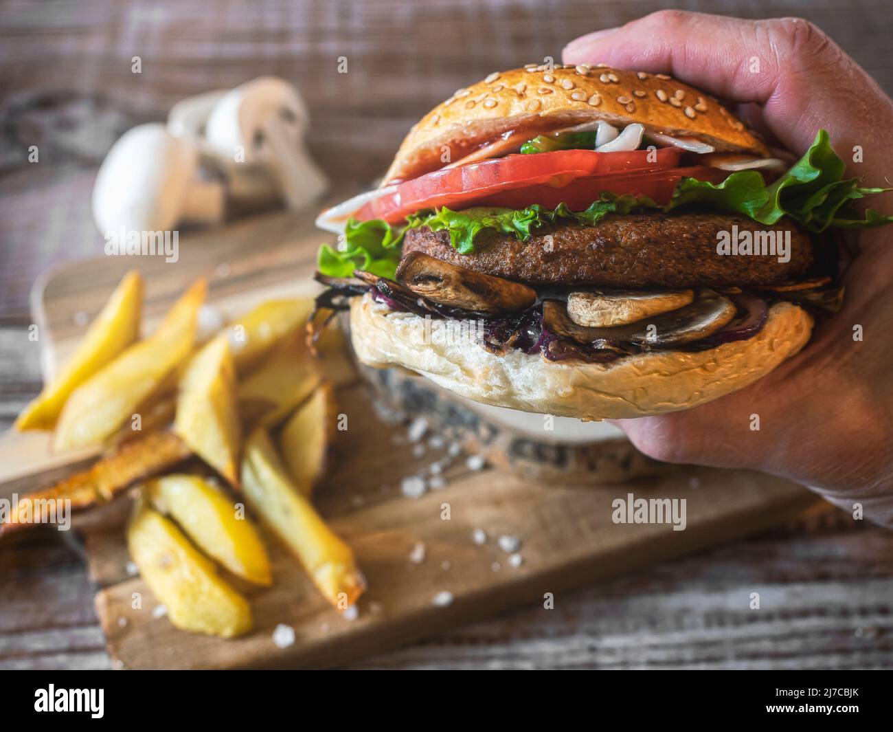 Hamburger mit Kichererbsen-Patty, Tomaten, Pilzen und Gemüse auf Roggenbrot. Eine Hand hält den Burger. Auf dem Boden Pommes und Pilze. Speicherplatz kopieren. Stockfoto