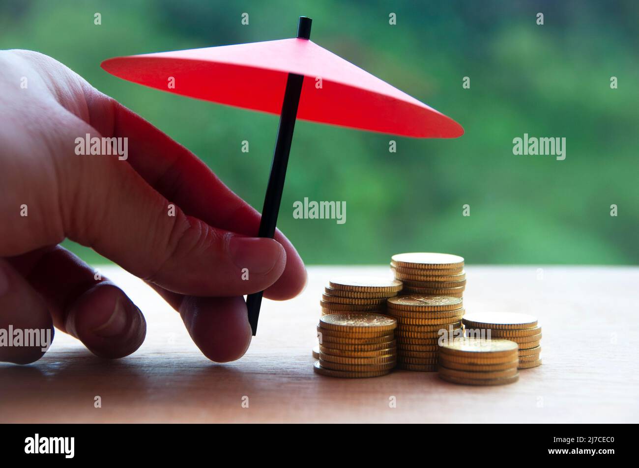 Hand hält kleinen roten Regenschirm über Haufen von Goldmünzen auf Holztisch. Nahaufnahme des Stapels von Goldmünzen mit der Hand, die den Regenschirm zum Schutz hält Stockfoto