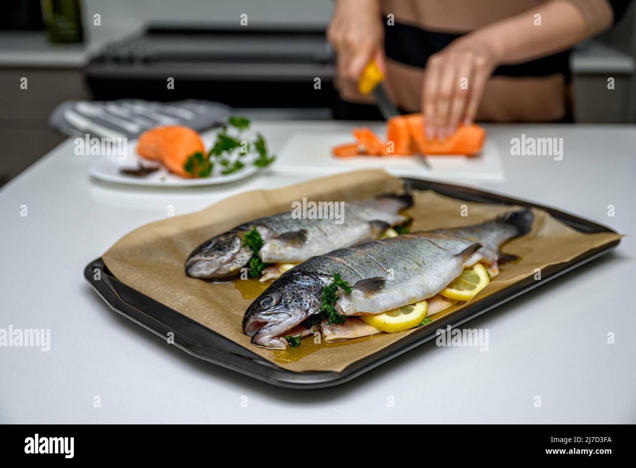 Kaukasische Frau in der Küche, die die Zutaten für ein gesundes Abendessen zubereitet. Sie stellte Regenbogenforelle mit Zitronenscheiben auf ein Backblech in einer Bratpfanne Stockfoto