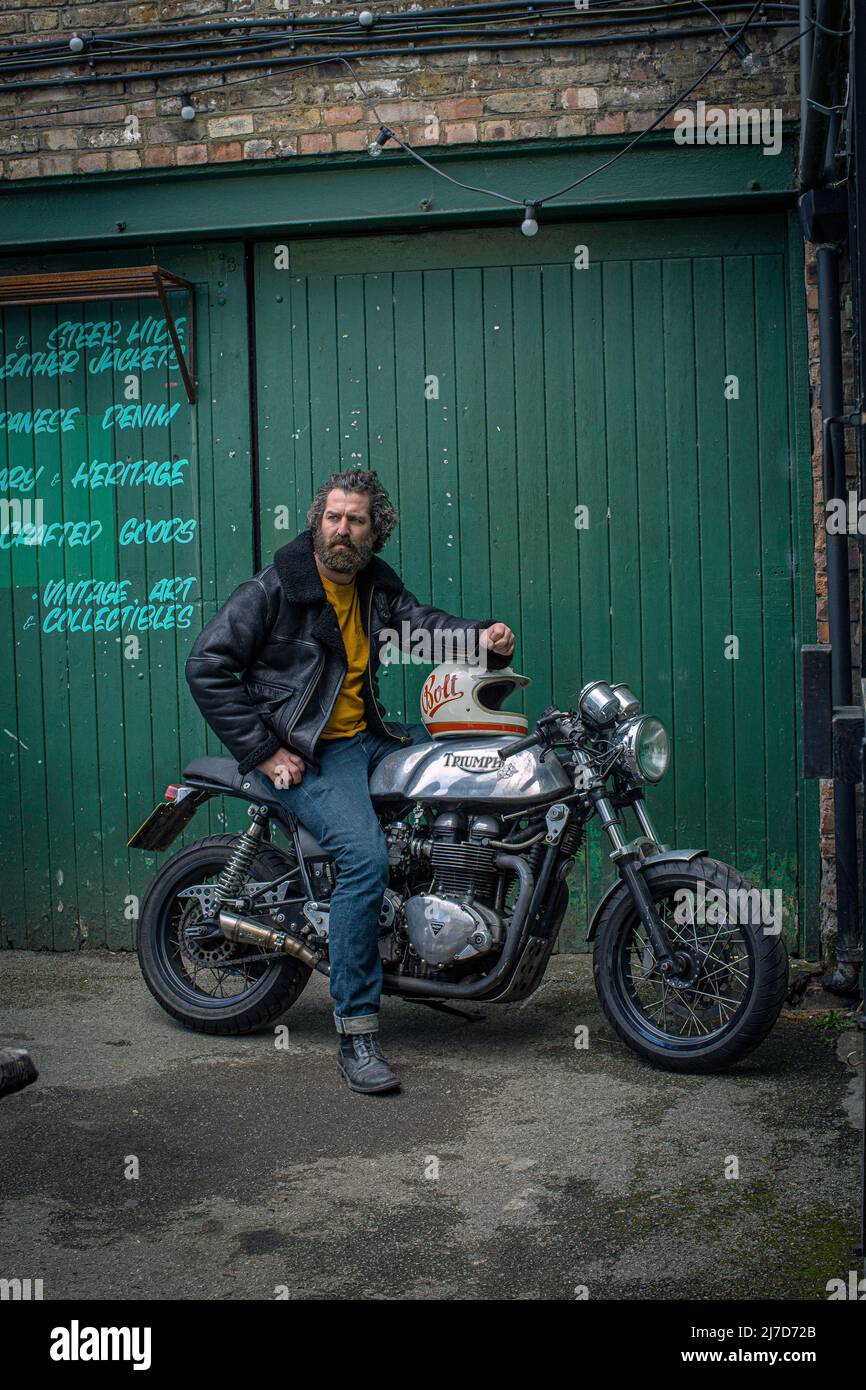 Männlich mit Bart trägt Lederjacke sitzt auf Cafe Racer Motorrad vor der Garage . Stockfoto
