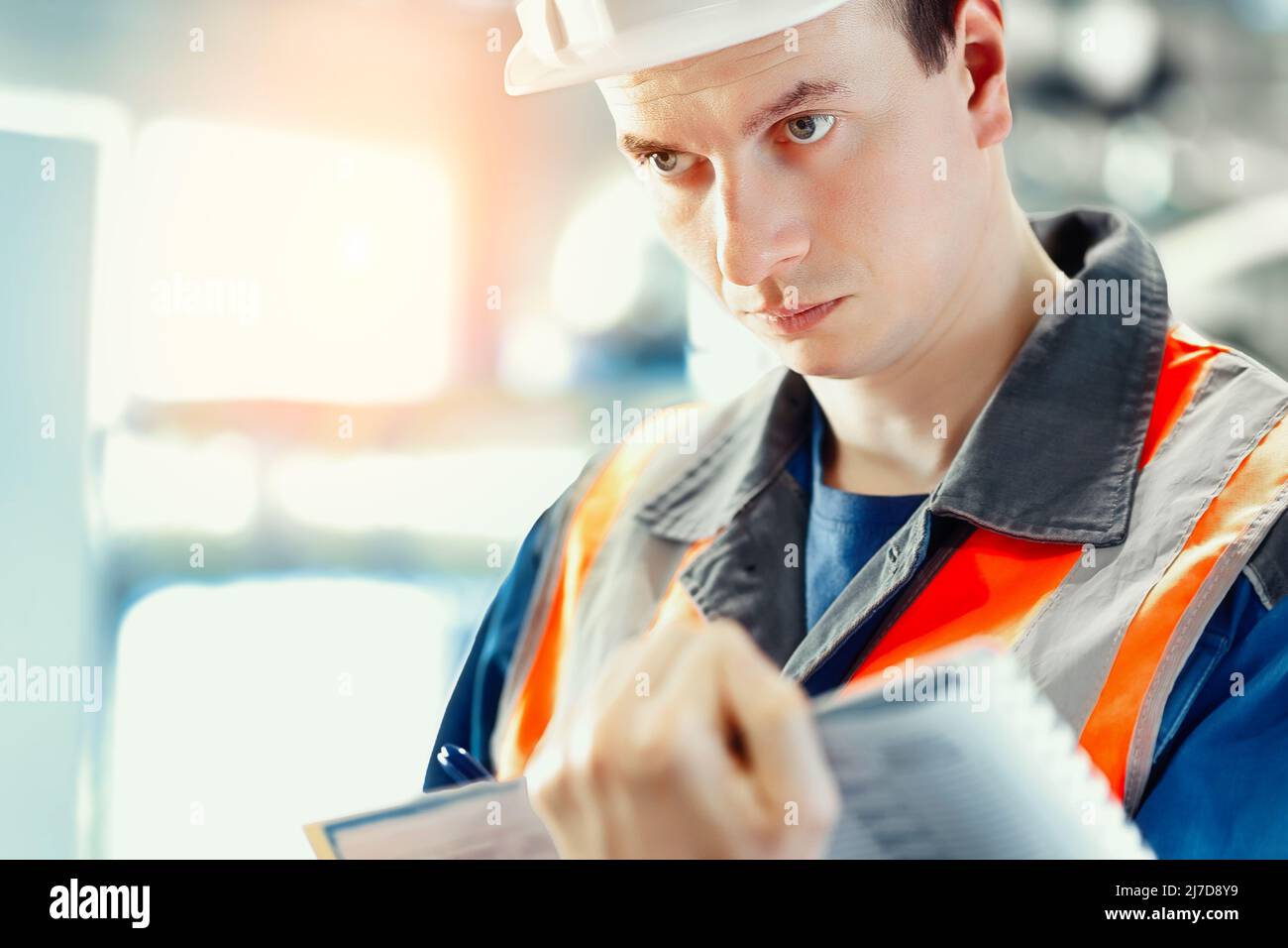 Der junge Inspektor oder Ingenieur in Helm und Bauweste führt Journaleinträge in der Produktionsanlage durch. Qualitätskontrolle der Arbeiten. Porträt eines kaukasischen Arbeiters im Zimmer. Stockfoto