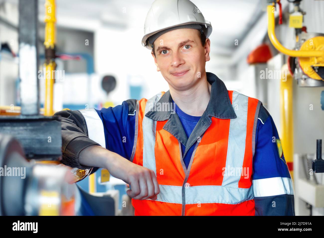 Junger positiver Ingenieur in Helm und Konstruktionsweste blickt in die Kamera und lächelt. Porträt eines jungen Schwerarbeiters im Raum mit unscharfem Hintergrund. Mitarbeiter in der Gasindustrie. Stockfoto