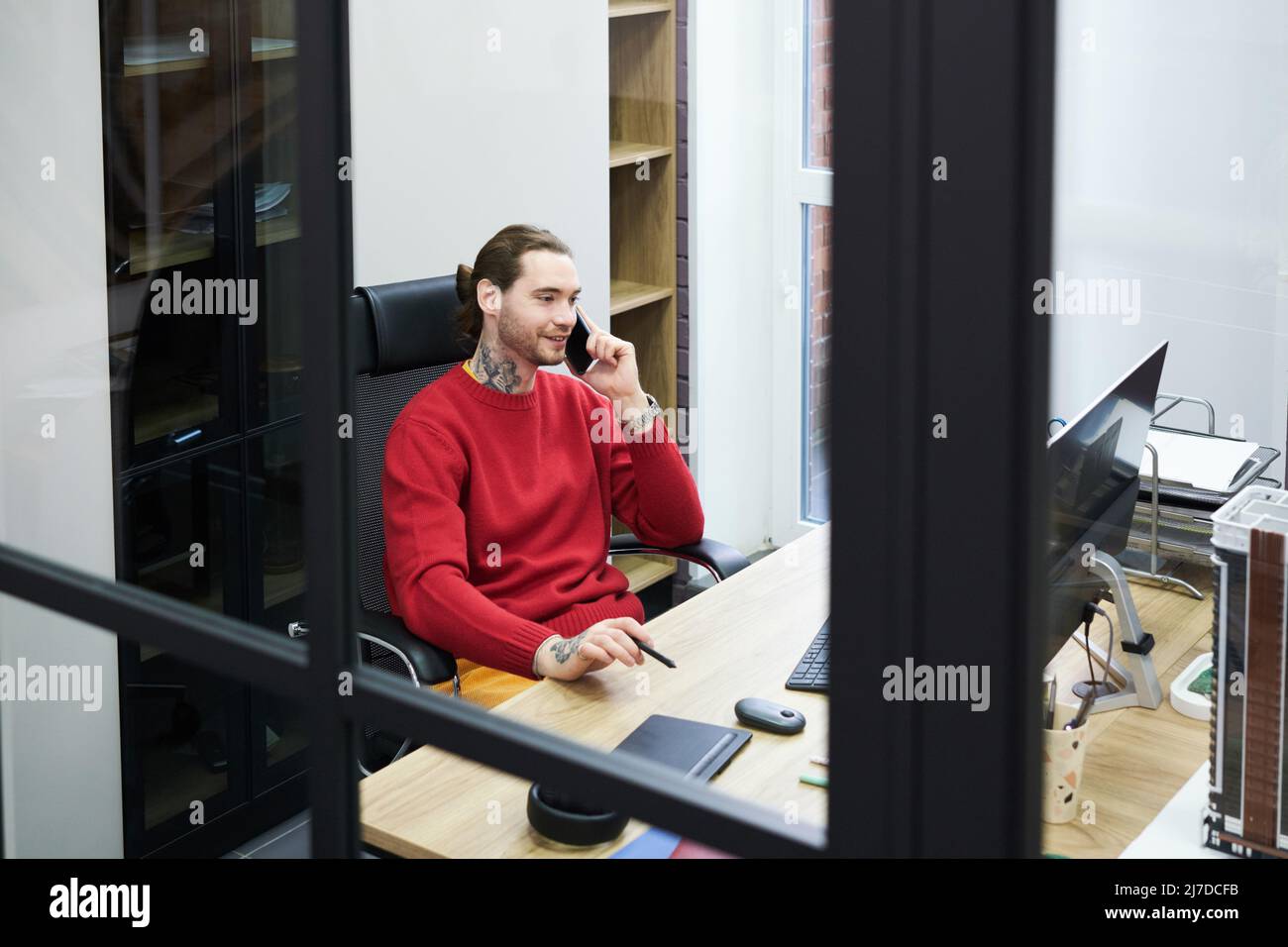 Lächelnder Manager sitzt am Tisch im Büro vor dem Computermonitor und führt Geschäftsgespräche auf dem Mobiltelefon Stockfoto