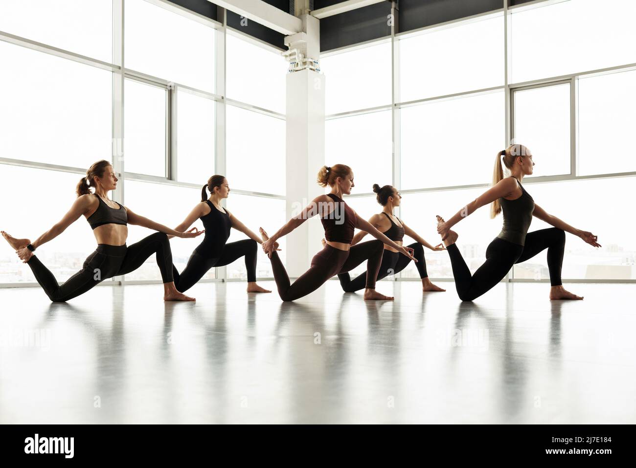 Eine Gruppe sportlicher Frauen in Schwarz steht auf einem Knie und dehnt das Bein, während sie den Körper mit Yoga-Übungen kontrolliert Stockfoto