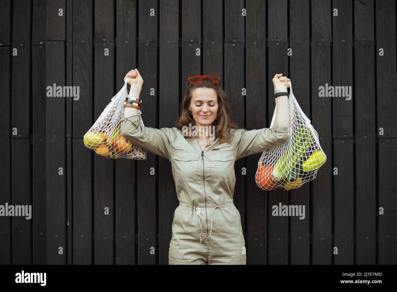 Lächelnde trendige Frau im Ganzen mit Saitenbeutel an der schwarzen Holzwand im Freien in der Stadt. Stockfoto