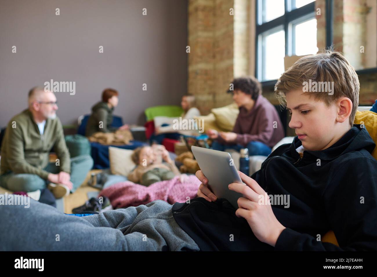 Jugendlicher Junge mit mobilen Gadgets, der sich die neuesten Nachrichten im Internet ansieht, während er sich auf dem Bett gegen Flüchtlinge ausruht, die miteinander reden Stockfoto