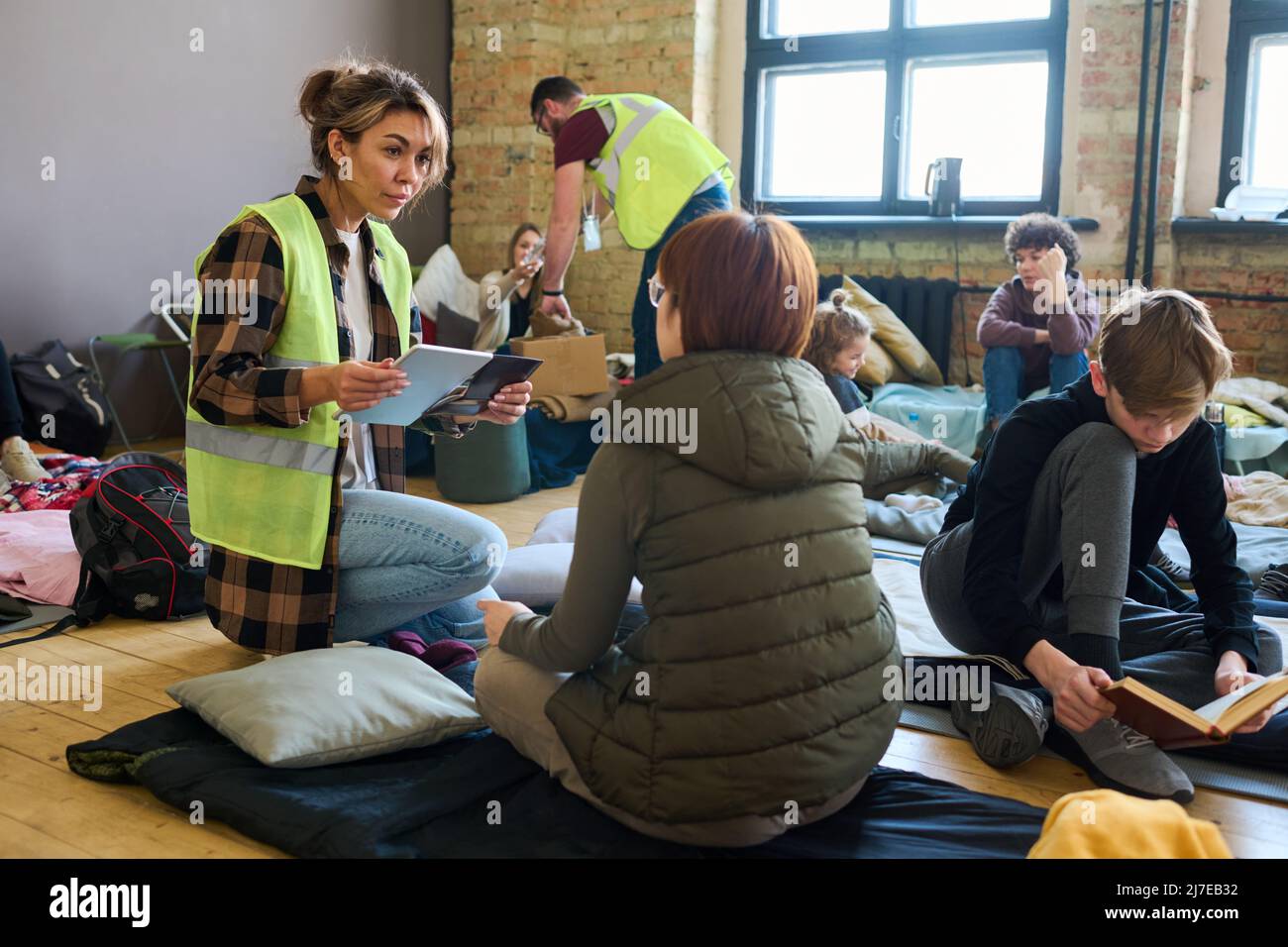 Weibliche Freiwillige mit mobilen Gadgets, die mit einem der Flüchtlinge kommunizieren, während sie auf Kniebeugen vor ihr im Unterschlupf für Migranten sitzt Stockfoto