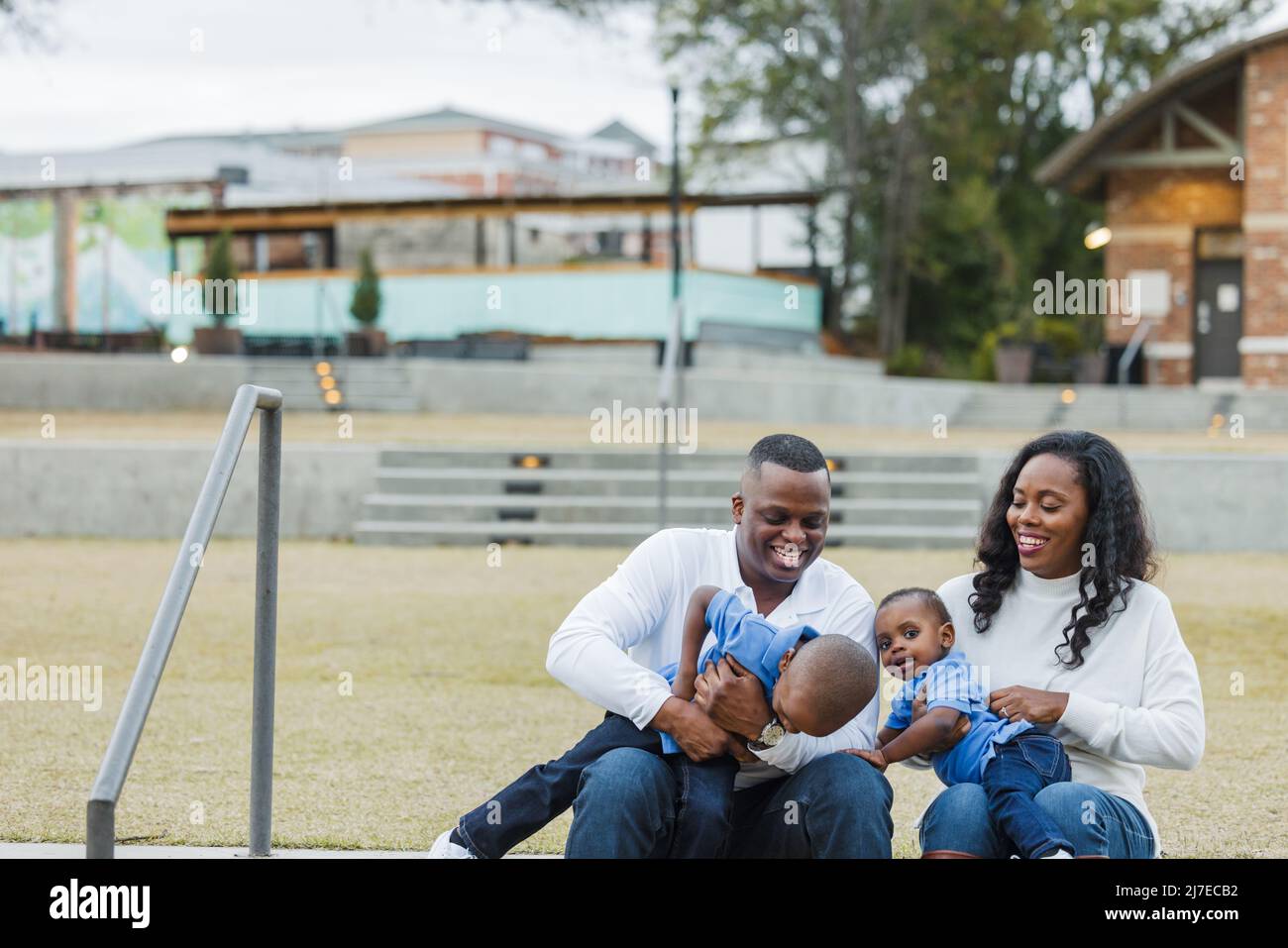 Eine wunderschöne afroamerikanische Mutter und ein wunderschöner Vater sitzen auf einer Treppe im Freien und kitzeln ihre beiden Söhne und alle lachen Stockfoto