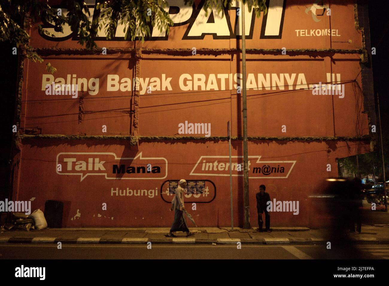 Menschen, die auf einem Bürgersteig vor einer Gebäudewand stehen und gehen, die mit einer Produktwerbung von Telkomsel, einem indonesischen Mobilfunkanbieter, in Palembang, Südsumatra, Indonesien, dekoriert ist. Stockfoto