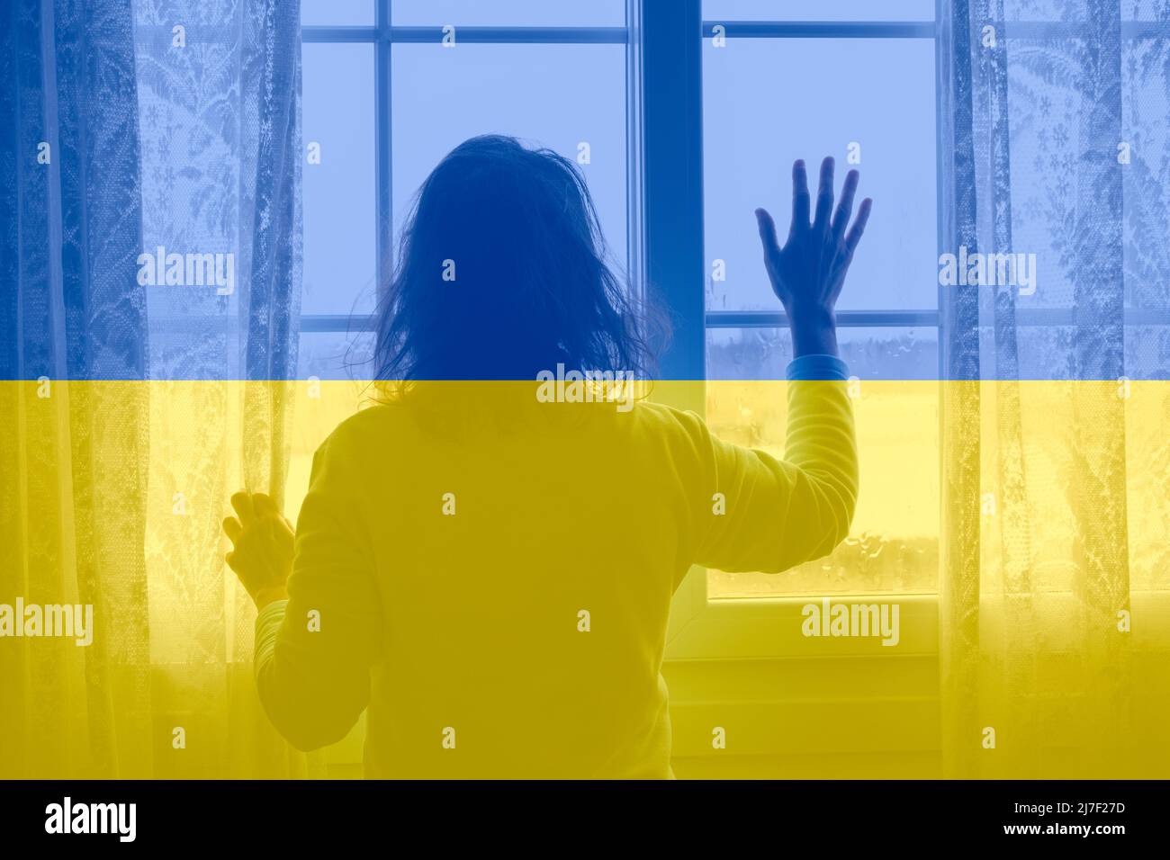Frau, die am regnerischen Tag aus dem Fenster schaute und die Flagge der Ukraine überdeckt hatte. Ukraine, Flüchtlinge, Flüchtlinge, Häuser für die Ukraine, psychische Gesundheit, Krieg, Russland Stockfoto