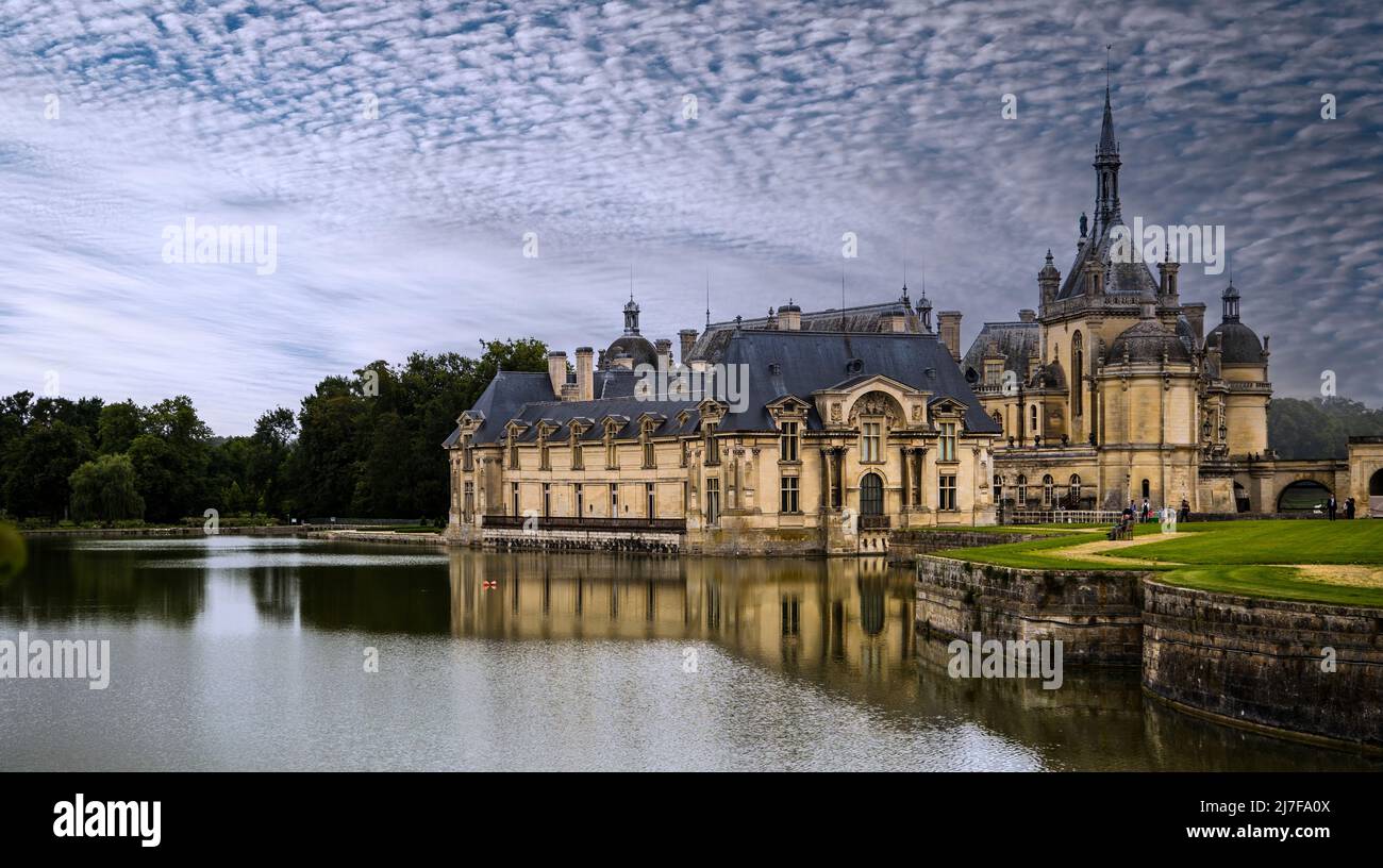 Panoramablick auf die Château de Chantilly, die sich im Wasser des Flusses mit hellen Wolken am Himmel widerspiegelt. Stockfoto