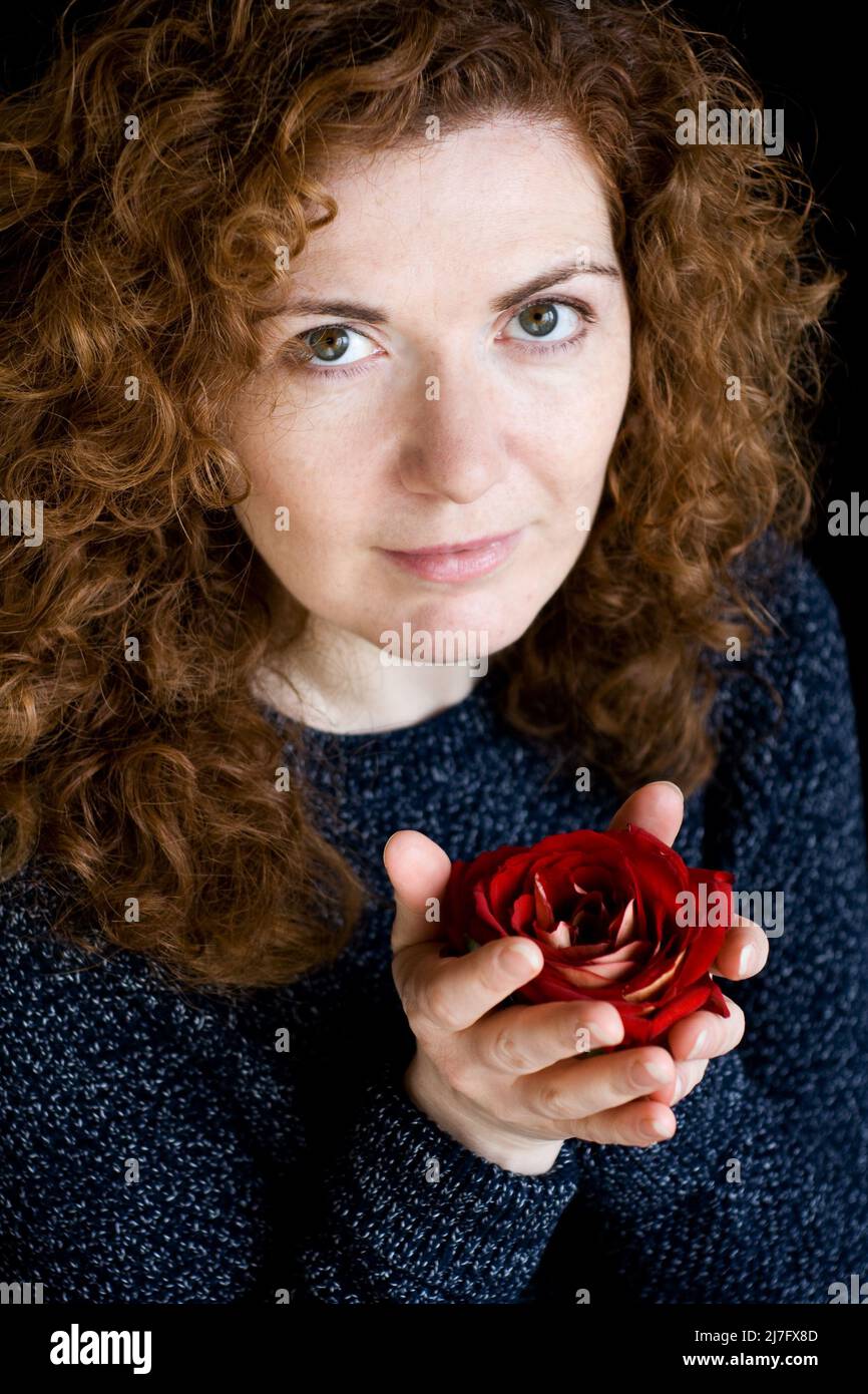 Porträt von schönen jungen Rotschopf mit lockigen roten Rose in der Hand auf einem dunklen Hintergrund Stockfoto