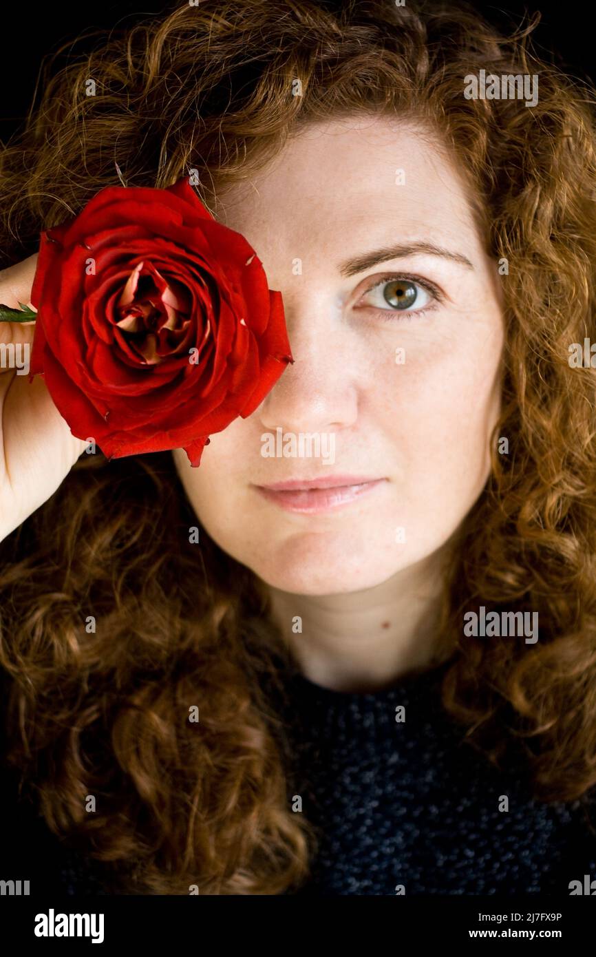 Porträt von schönen jungen Rotschopf mit lockigen roten Rose in der Hand auf einem dunklen Hintergrund Stockfoto