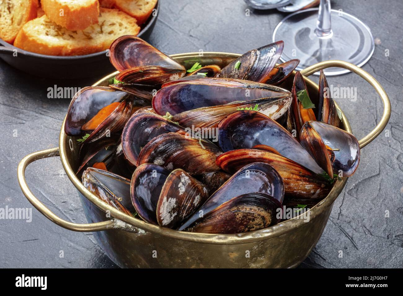 Gekochte Muscheln in einer Kupferpfanne, belgische Küche in einem Restaurant, mit geröstetem Brot und einem Glas Wein im Hintergrund Stockfoto