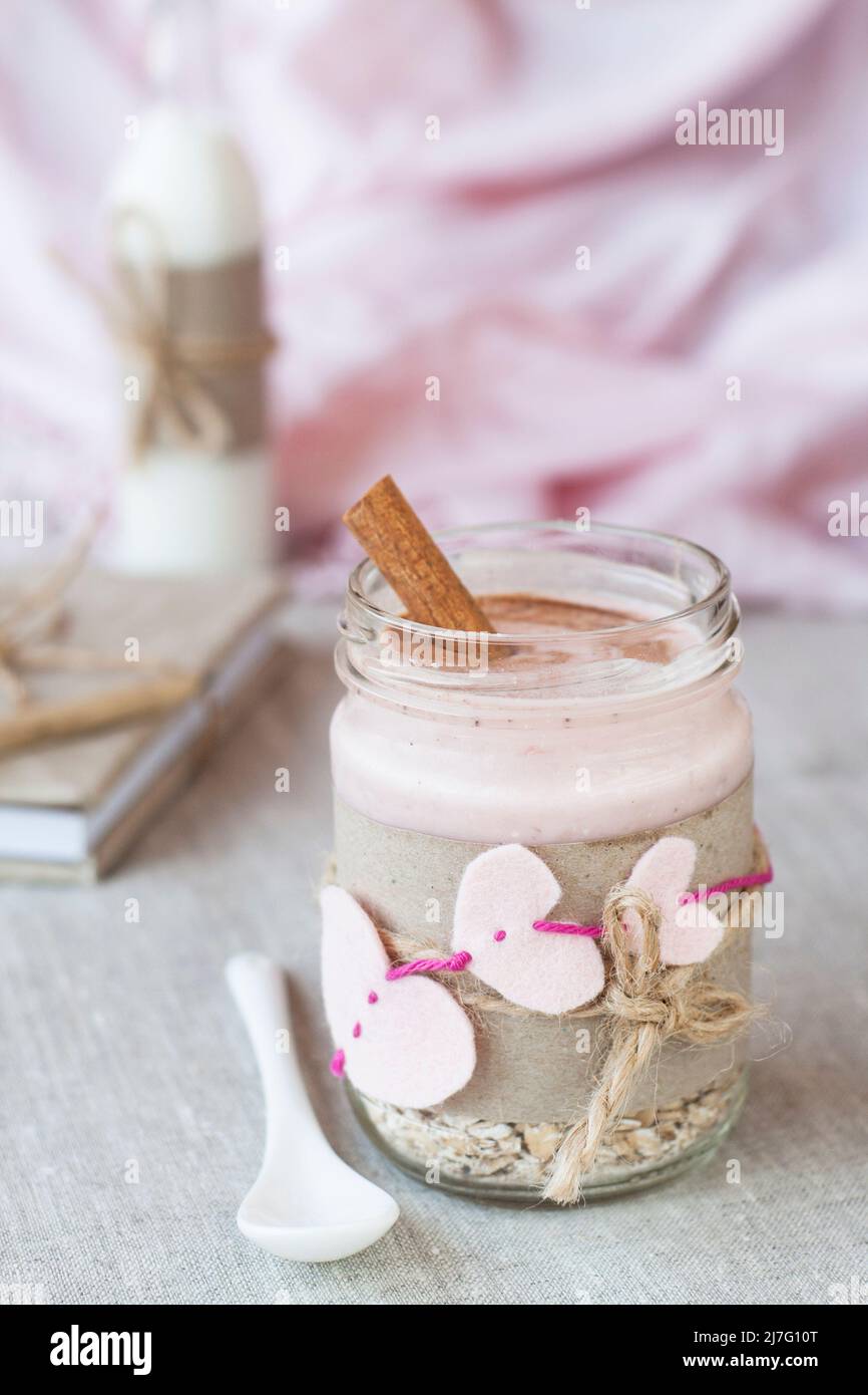 Haferflocken, gewürzt mit Joghurt, bestreut mit Zimt in einem Glas. Als nächstes kommt eine Flasche Milch. Als nächstes gibt es ein Bastelheft und eine rosa Serviette. Stockfoto