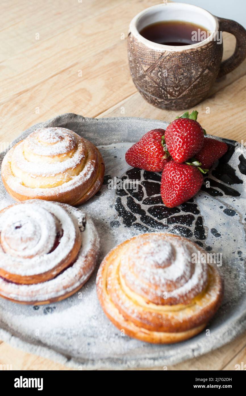 Köstliche Brötchen werden mit Zuckerpulver bestreut und rote, saftige Erdbeeren liegen auf einem hellen, handgefertigten Teller auf hellem Holzhintergrund. Als nächstes ist eine Tasse o Stockfoto